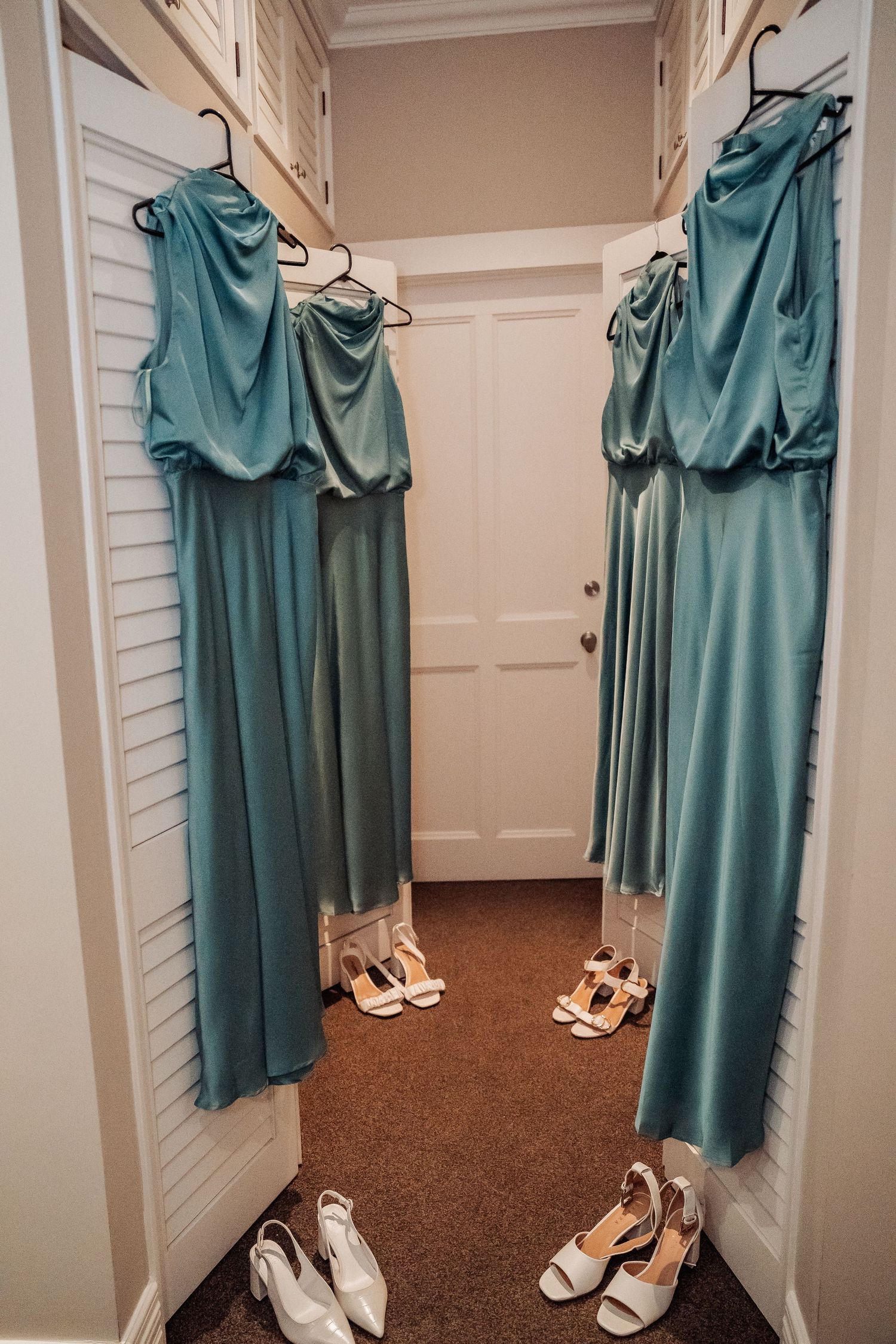 Turquoise bridesmaid dresses hanging in a closet with matching white heels arranged on the brown carpeted floor.