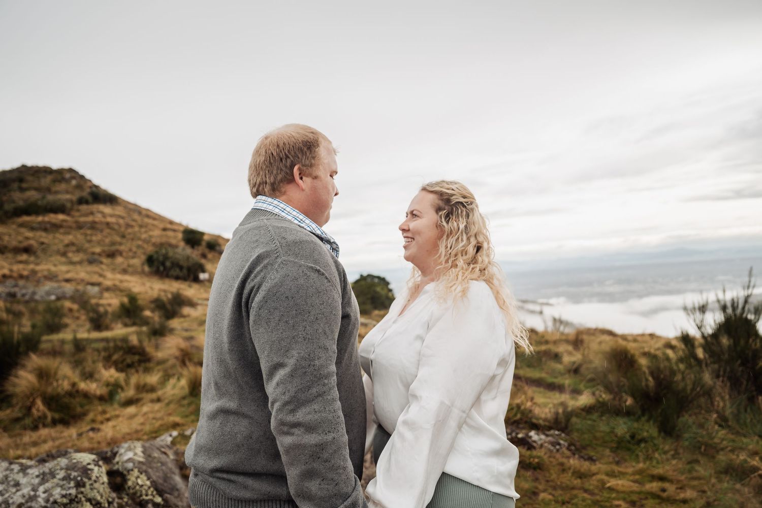 A couple stands close together on a hilltop overlooking a dramatic coastal landscape on an overcast day.