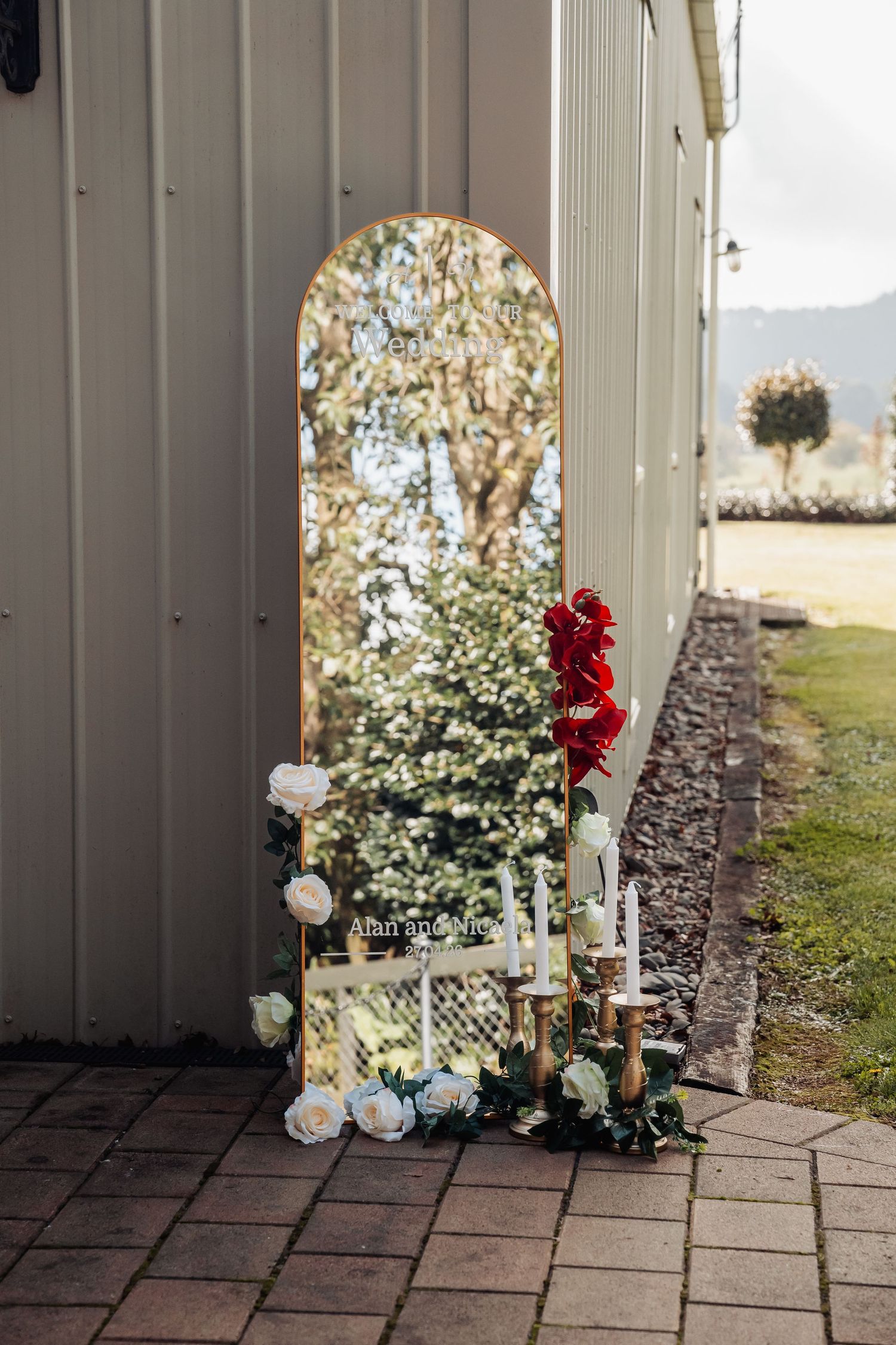 Elegant wedding mirror with gold arch frame, white and red floral arrangements and candles outdoors.