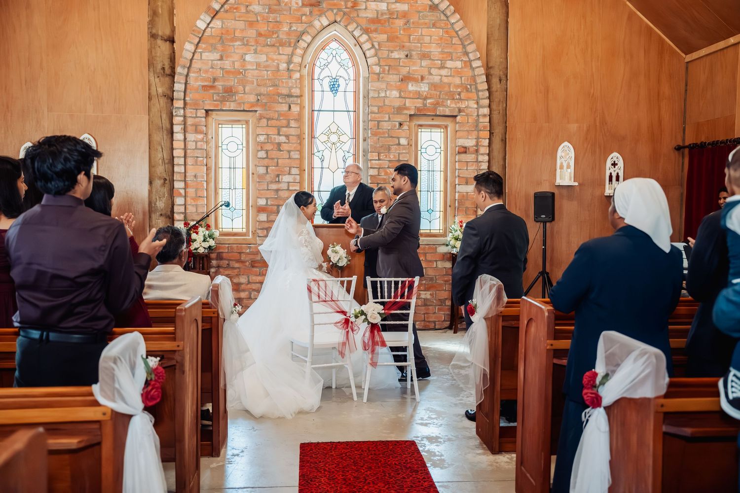 Bride and groom exchange vows at altar in rustic brick chapel with red rose petal aisle.