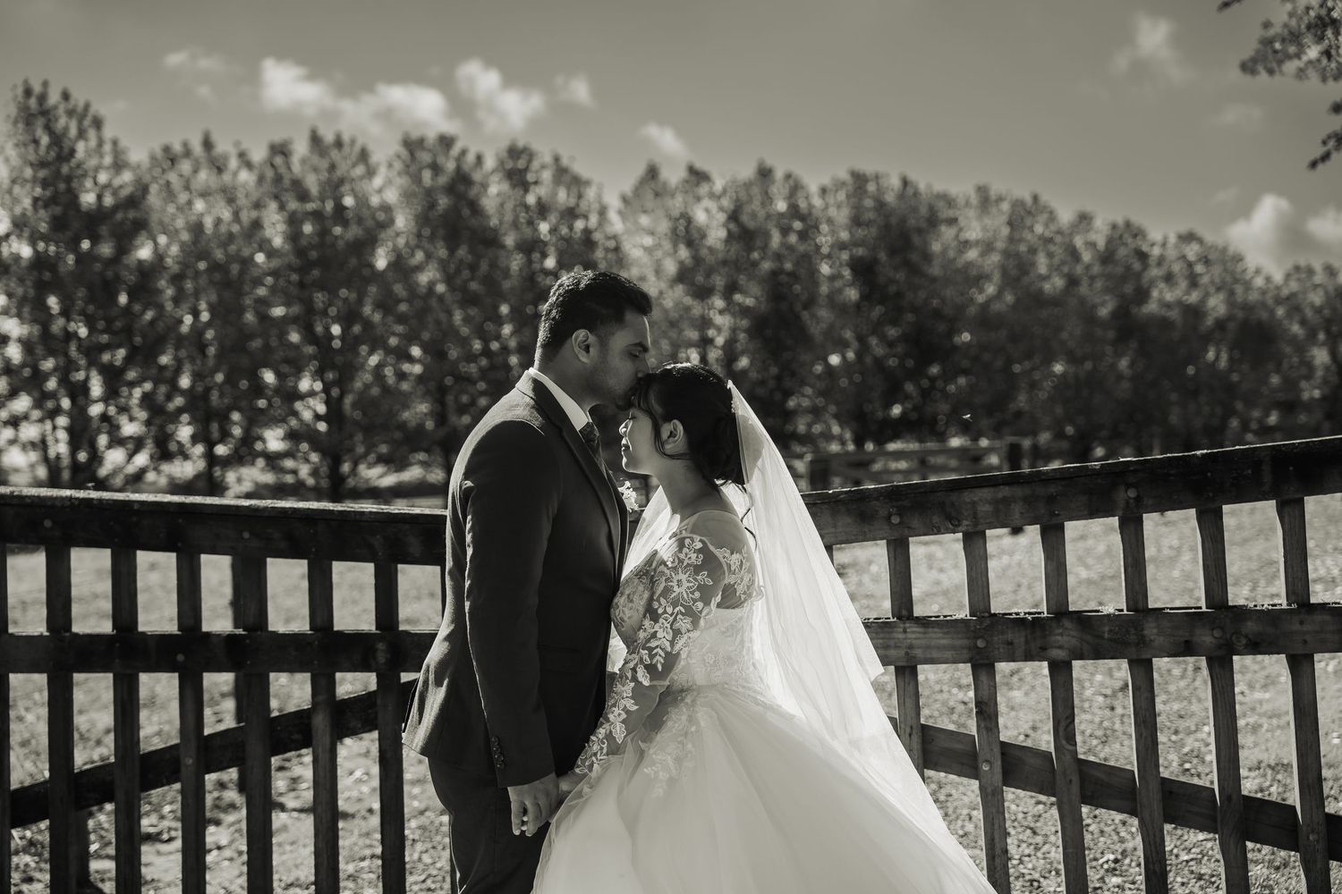 Black and white romantic photo of bride and groom sharing a tender moment by a wooden fence.