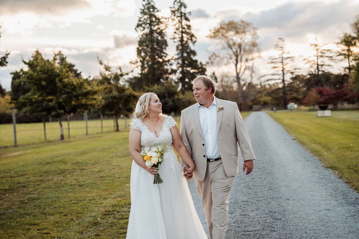 Bride and groom walk hand in hand down gravel path at sunset holding yellow bouquet.