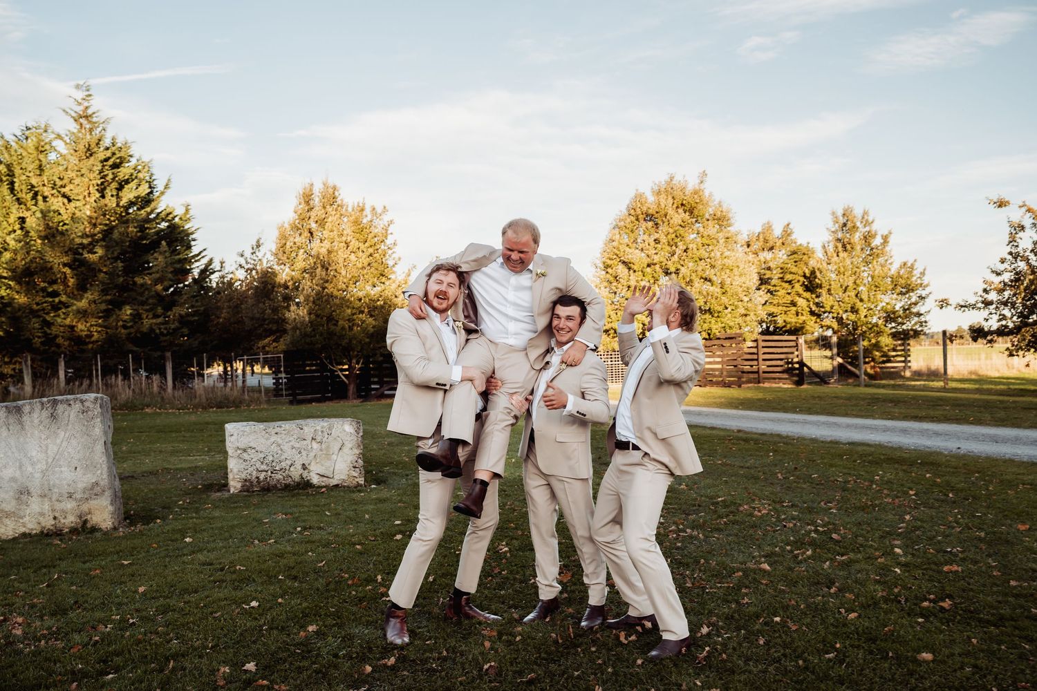 Joyful wedding party in tan suits celebrate outdoors lifting someone in the air.