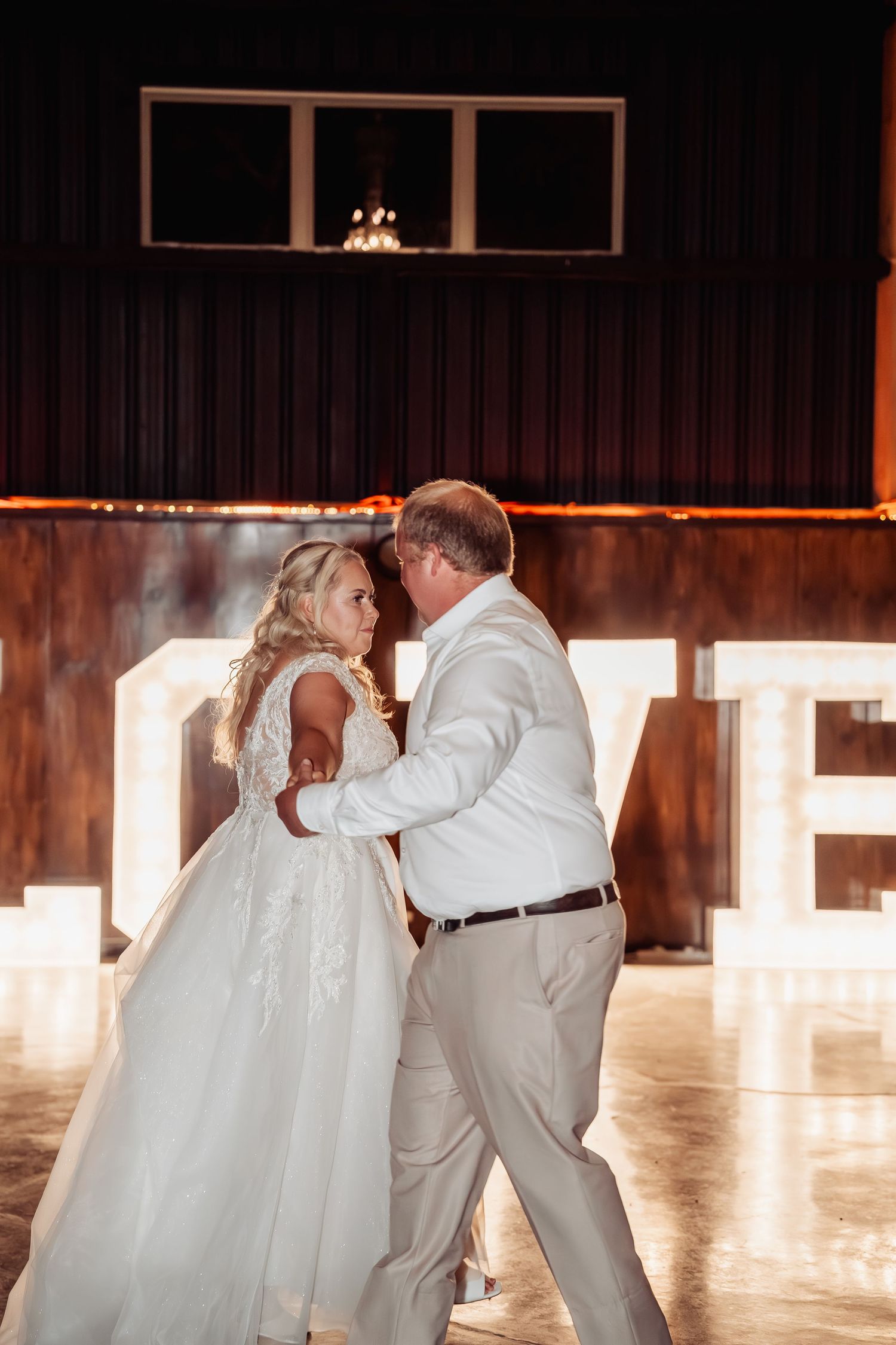 Bride and groom share first dance in front of illuminated LOVE letters at elegant indoor wedding reception.