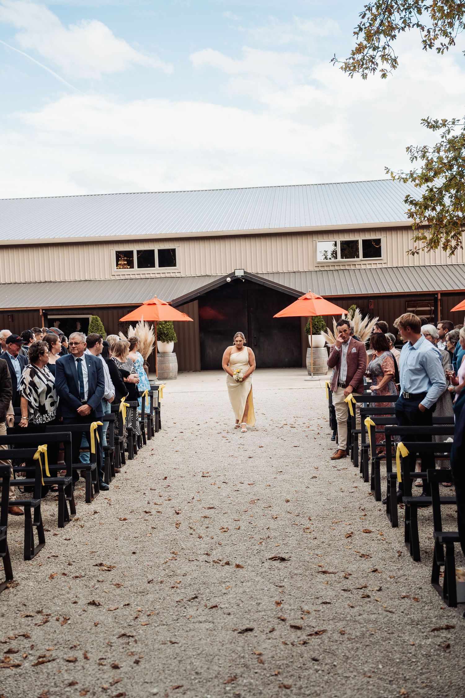 Bridesmaid in gold dress walking down outdoor gravel aisle at barn wedding venue.