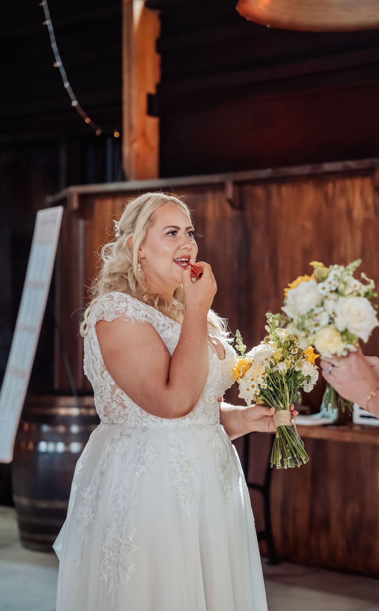 Blonde bride in lace wedding gown holding yellow and white bouquet at rustic barn venue.