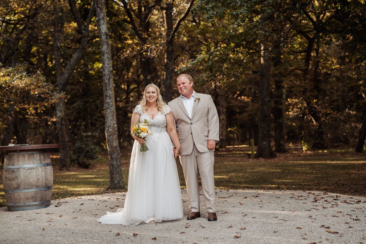 Bride in white gown holding yellow bouquet and groom in tan suit posing outdoors among autumn trees.