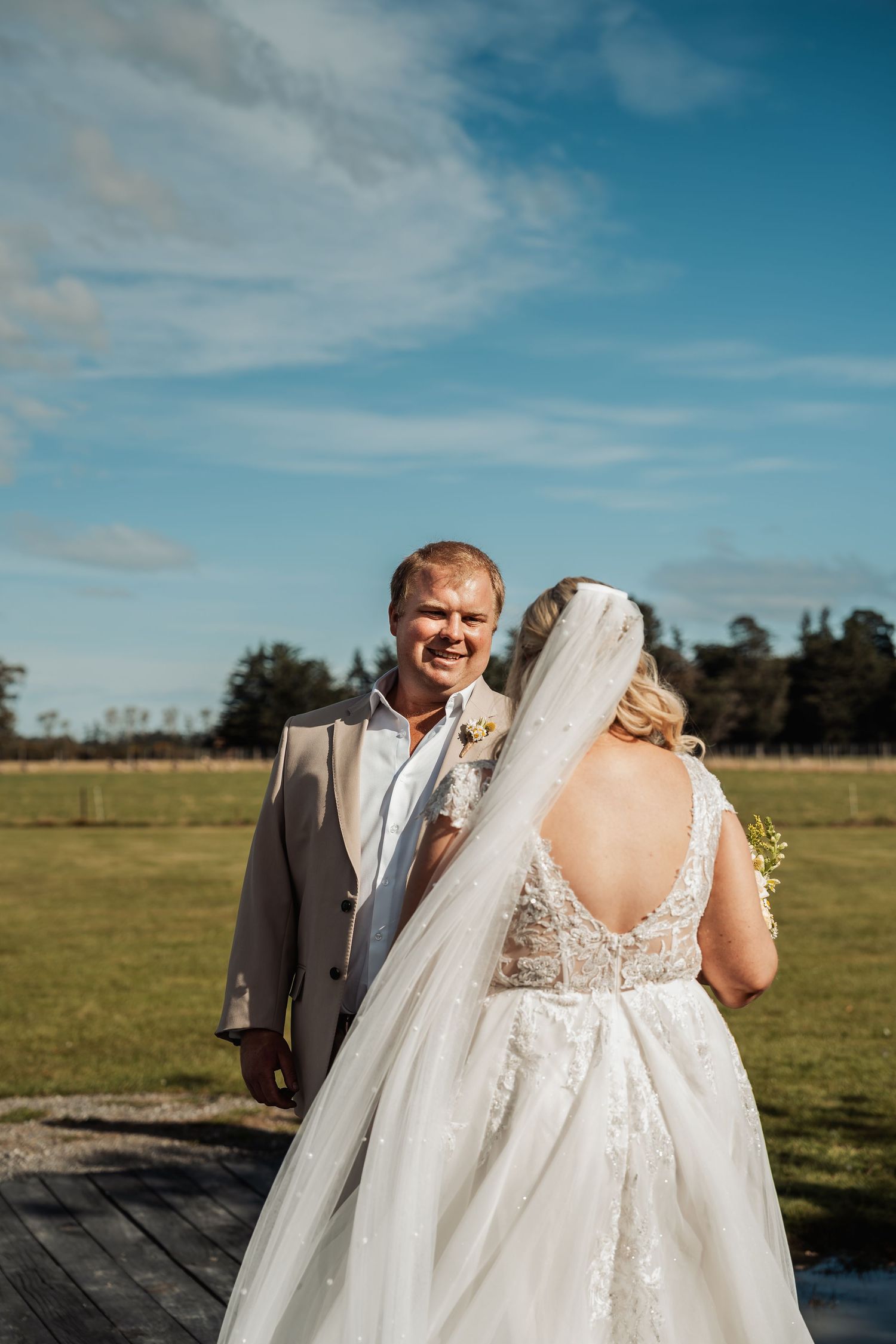 Smiling groom in tan suit turns to face bride in lace wedding dress outdoors.