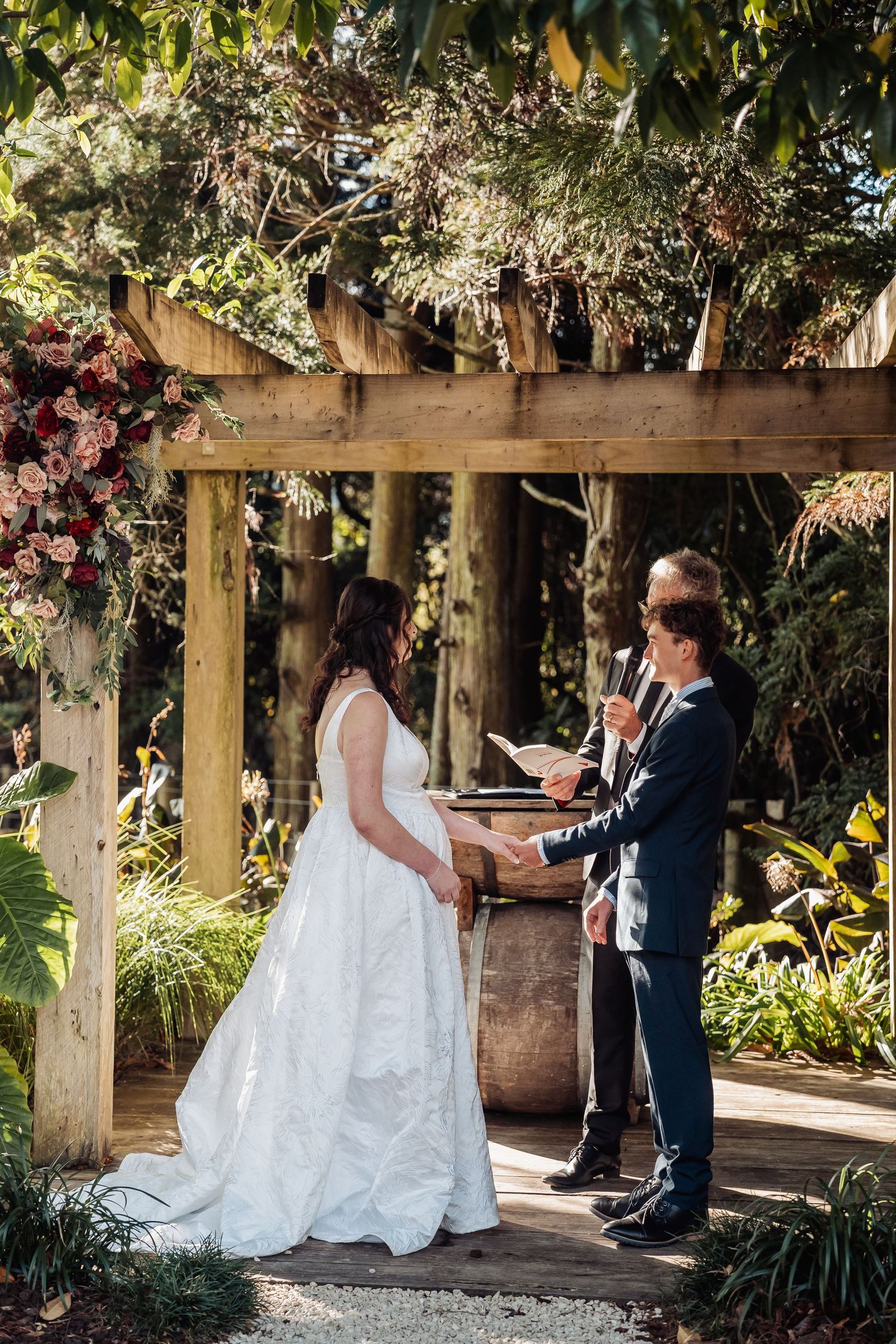 Bride and groom exchanging vows under floral wooden pergola in lush garden.