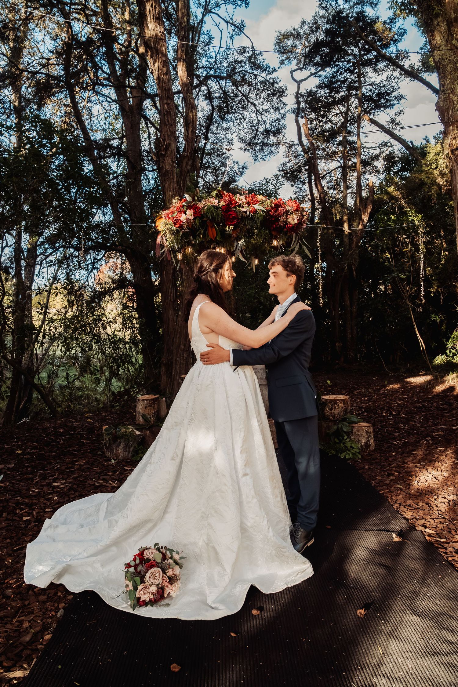 Bride and groom embracing under floral arch in sunlit forest wedding ceremony.