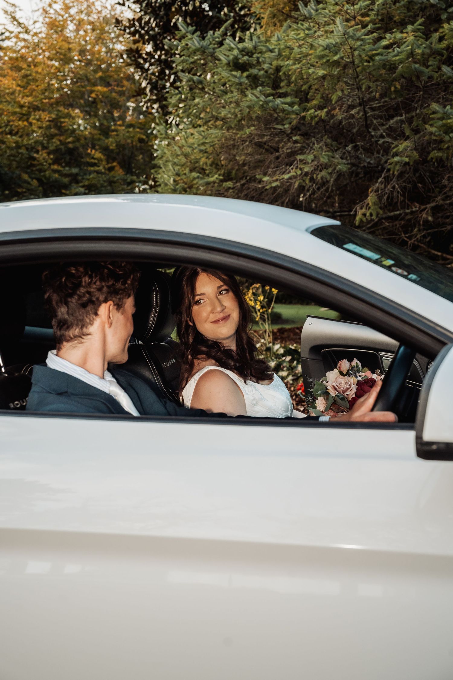 Bride holding a bouquet smiles at the groom while seated inside a white sports car surrounded by green trees.