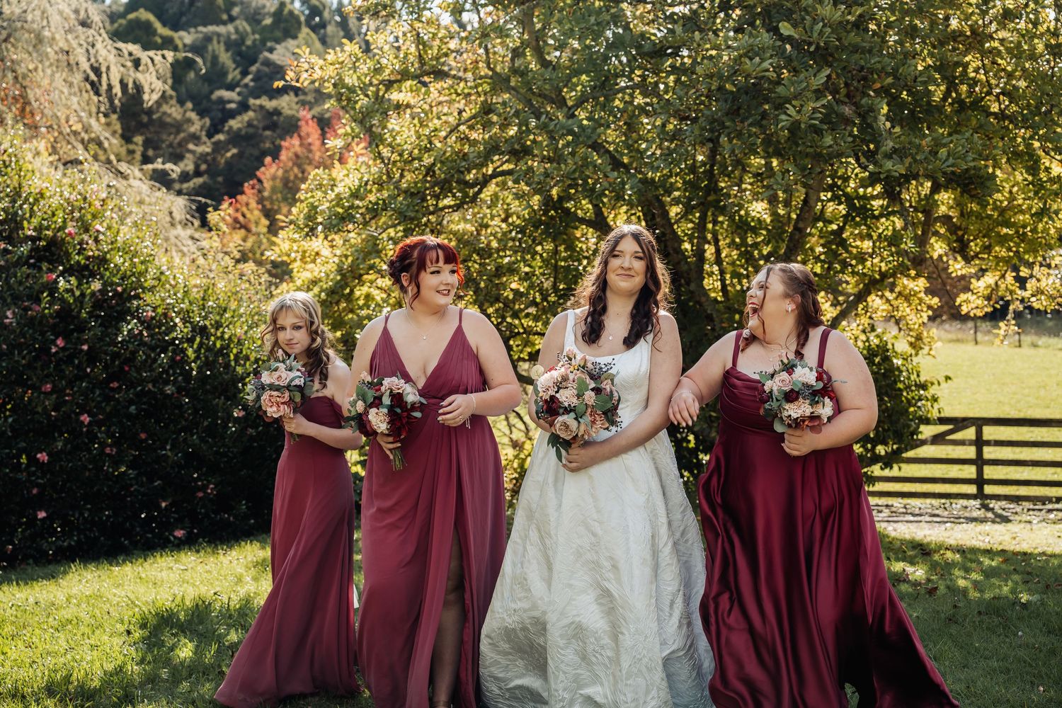 Bride in white gown walks with three bridesmaids in burgundy dresses, holding bouquets outdoors on a sunny autumn day.