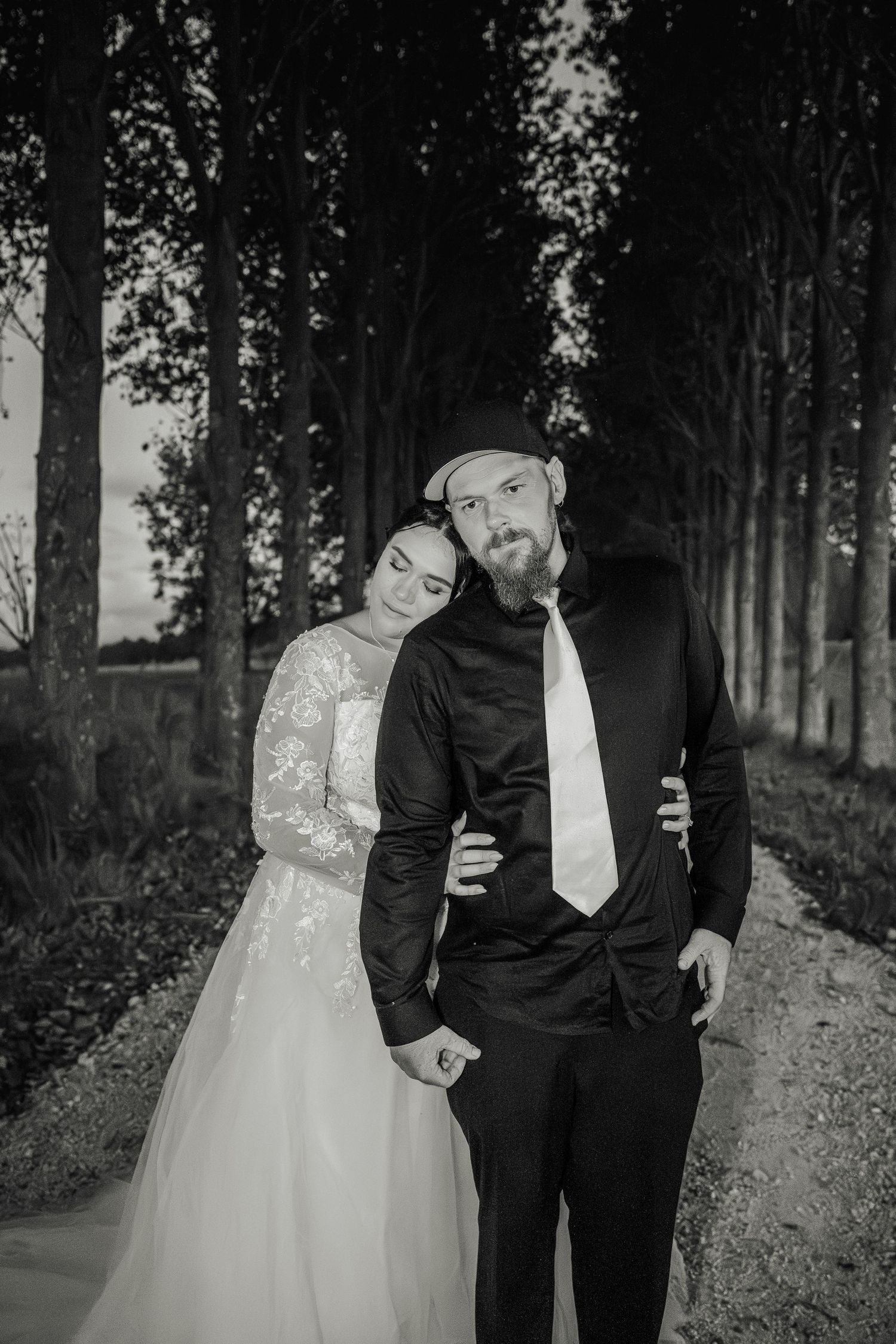 Black and white portrait of couple in formal attire standing together on tree-lined path.