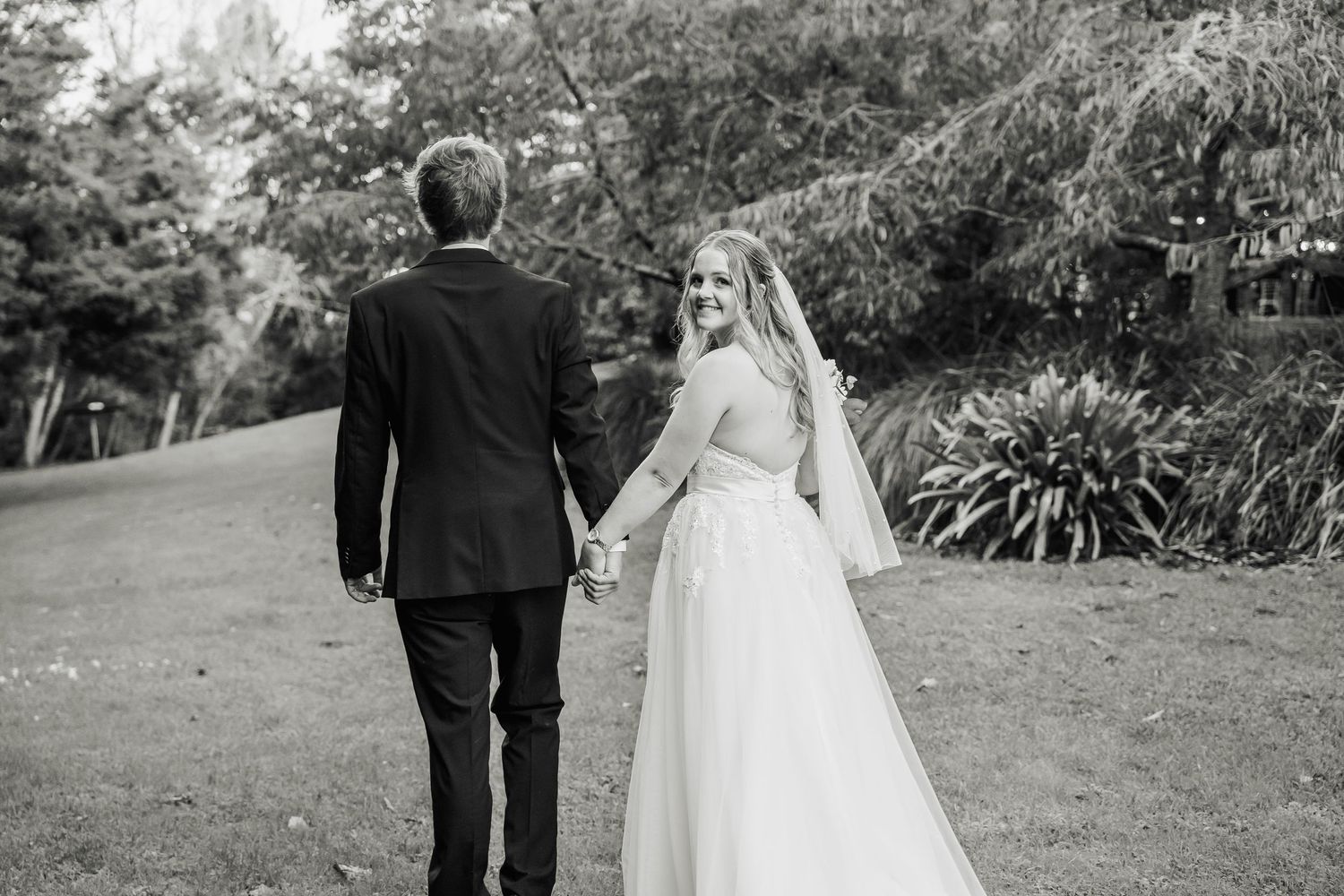A black and white wedding photograph captures a couple walking hand in hand through a garden setting.