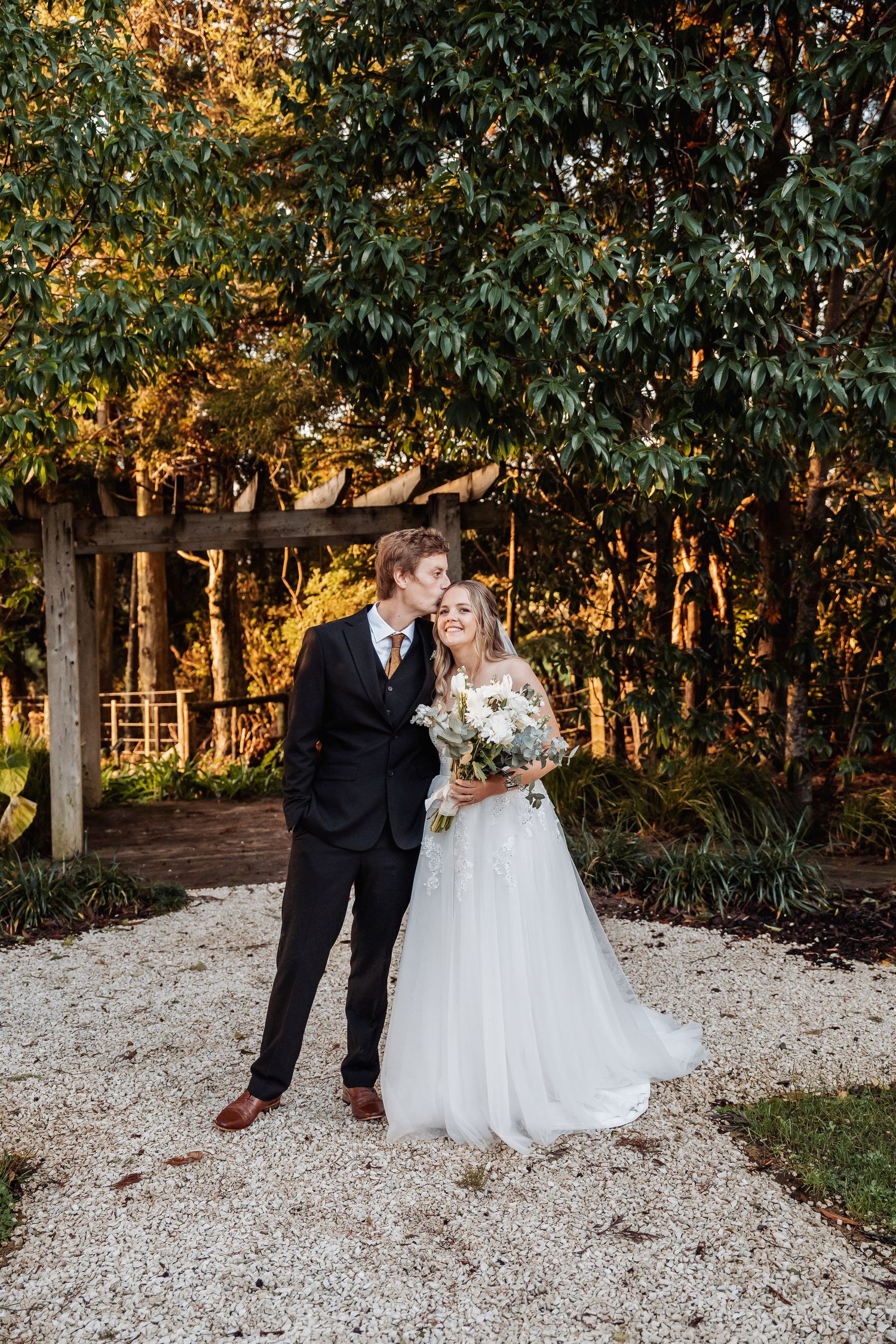 A romantic outdoor wedding photo on a gravel path with sunlit trees in the background during golden hour.