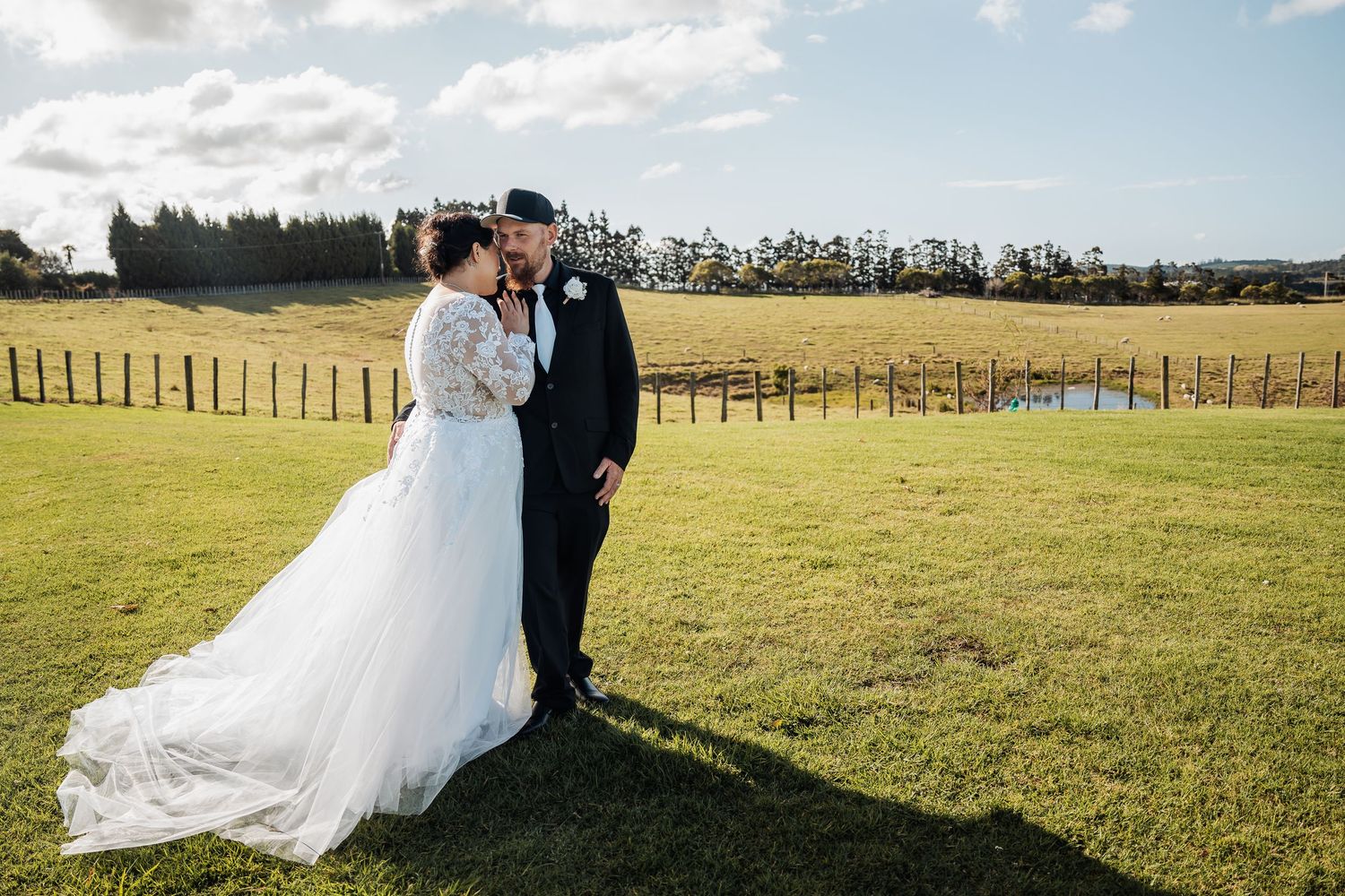 Newlyweds stand together on rolling green hills with wooden fences and trees in the countryside on their wedding day.