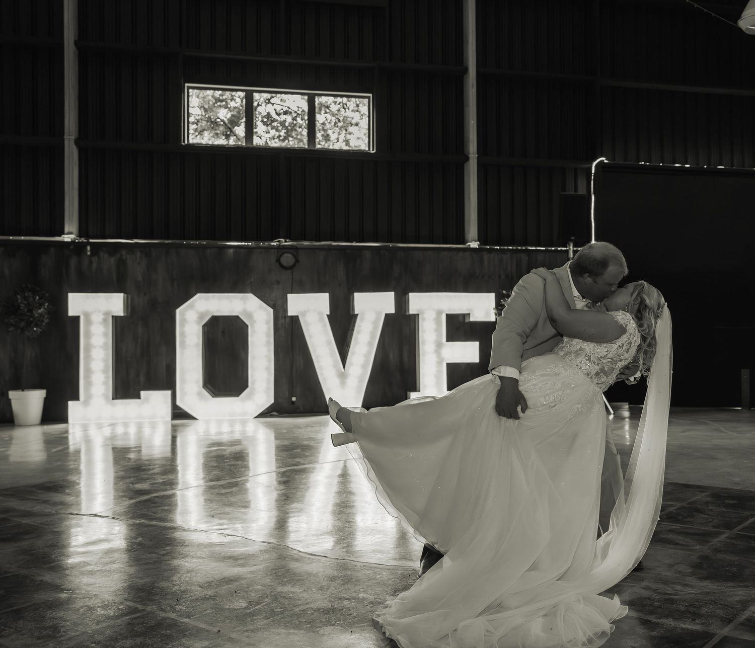 Black and white photo of a bride and groom sharing a romantic dip kiss in front of illuminated LOVE letters.
