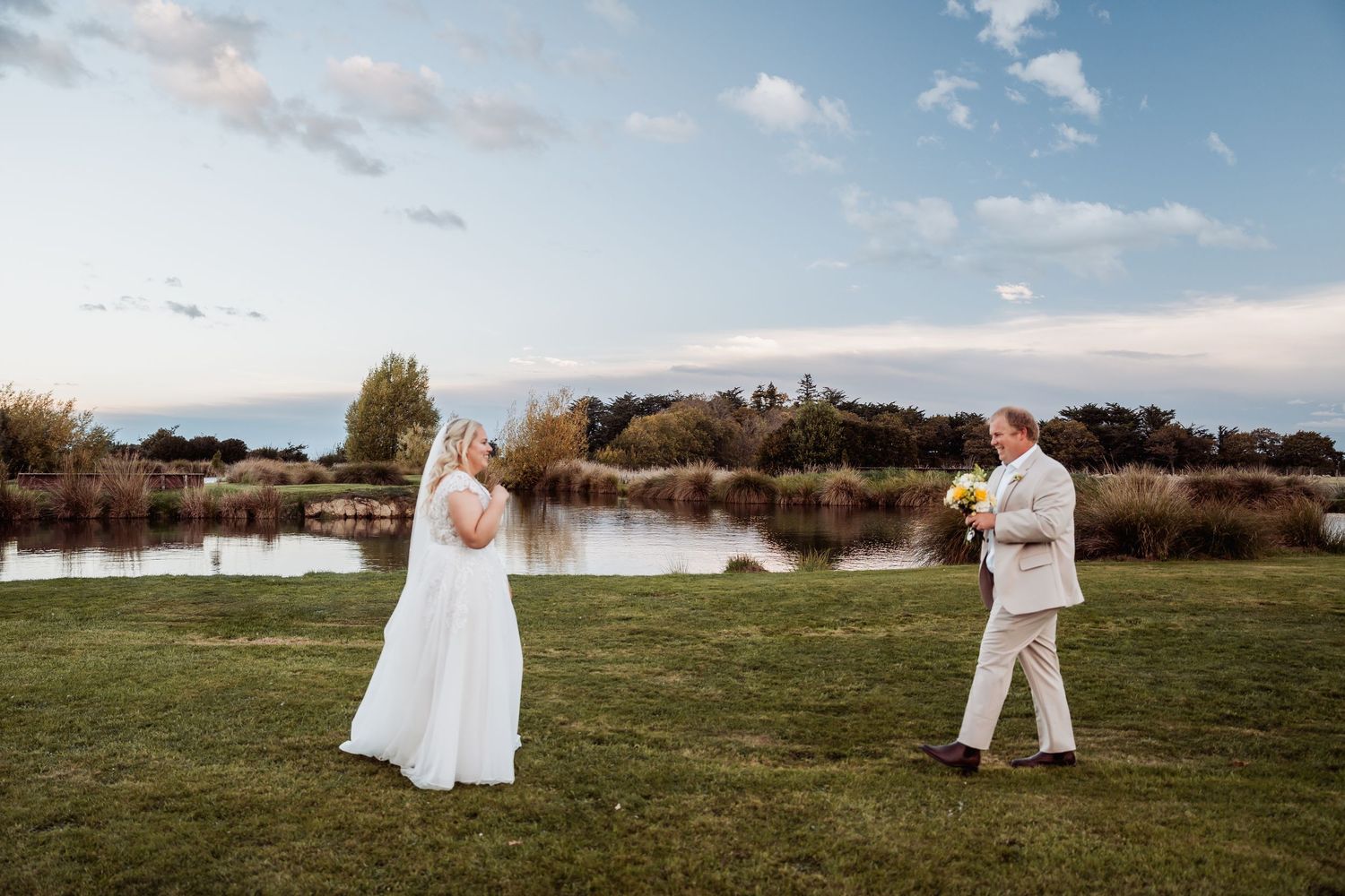 Bride in white gown and groom in tan suit share a first look moment by a scenic pond.