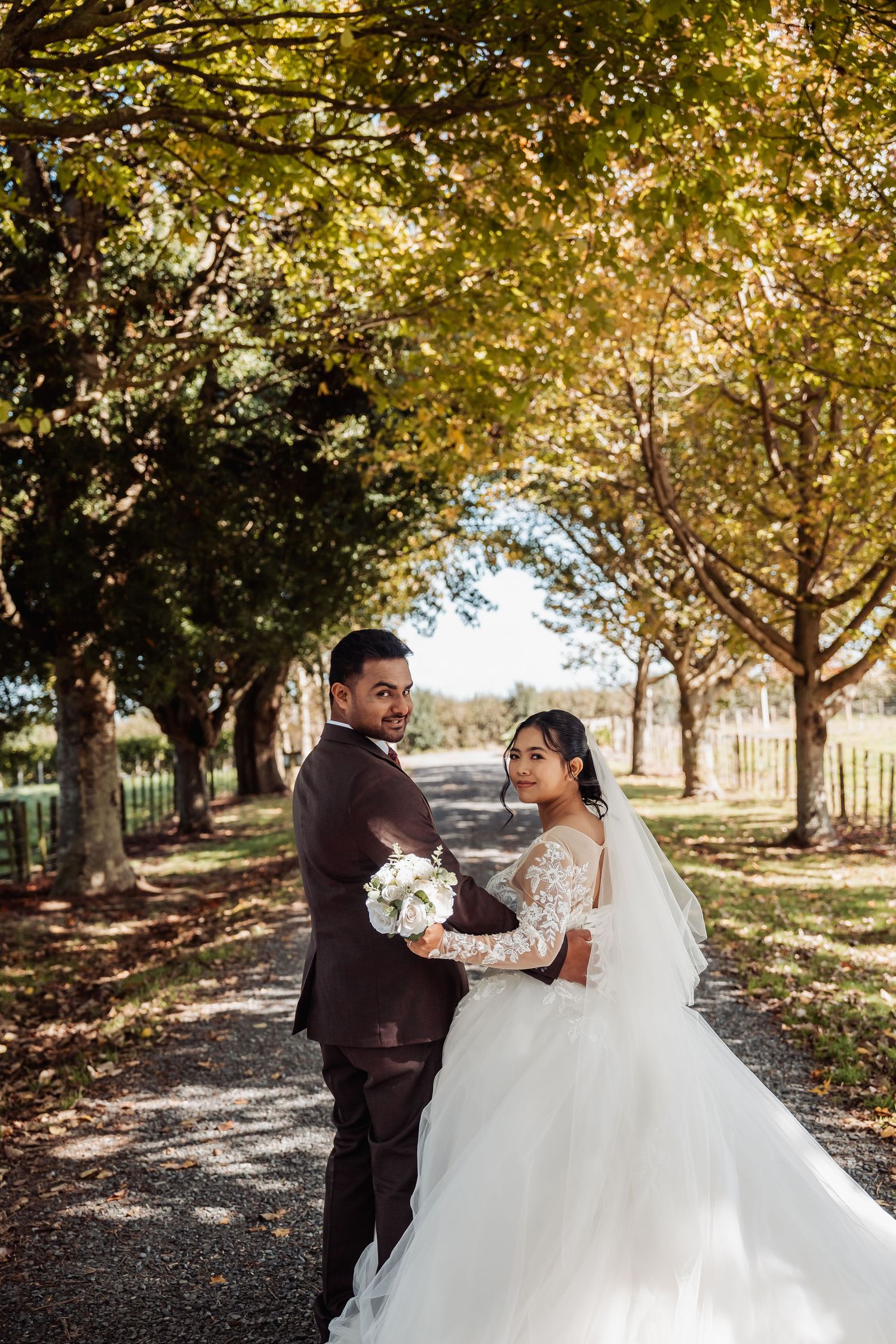 Bride and groom posing on a tree-lined path in autumn, bride holding white bouquet in elegant lace wedding gown.