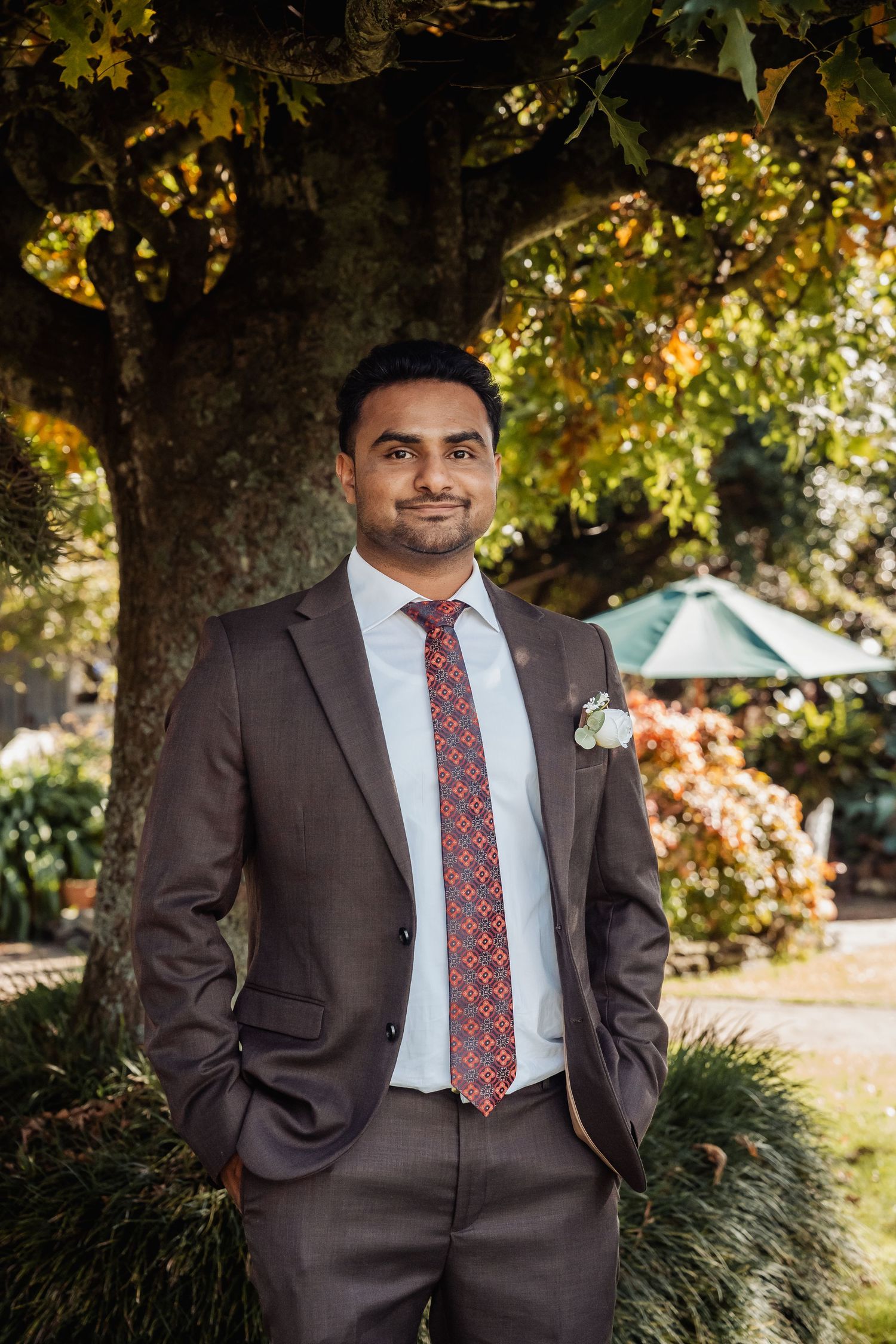 Groom in brown suit with floral tie and boutonniere poses outdoors under autumn tree at wedding.