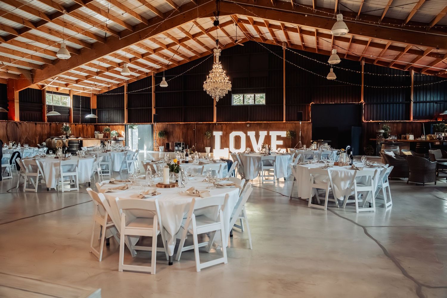 Rustic barn wedding reception with white round tables, chandelier, and illuminated LOVE sign backdrop.