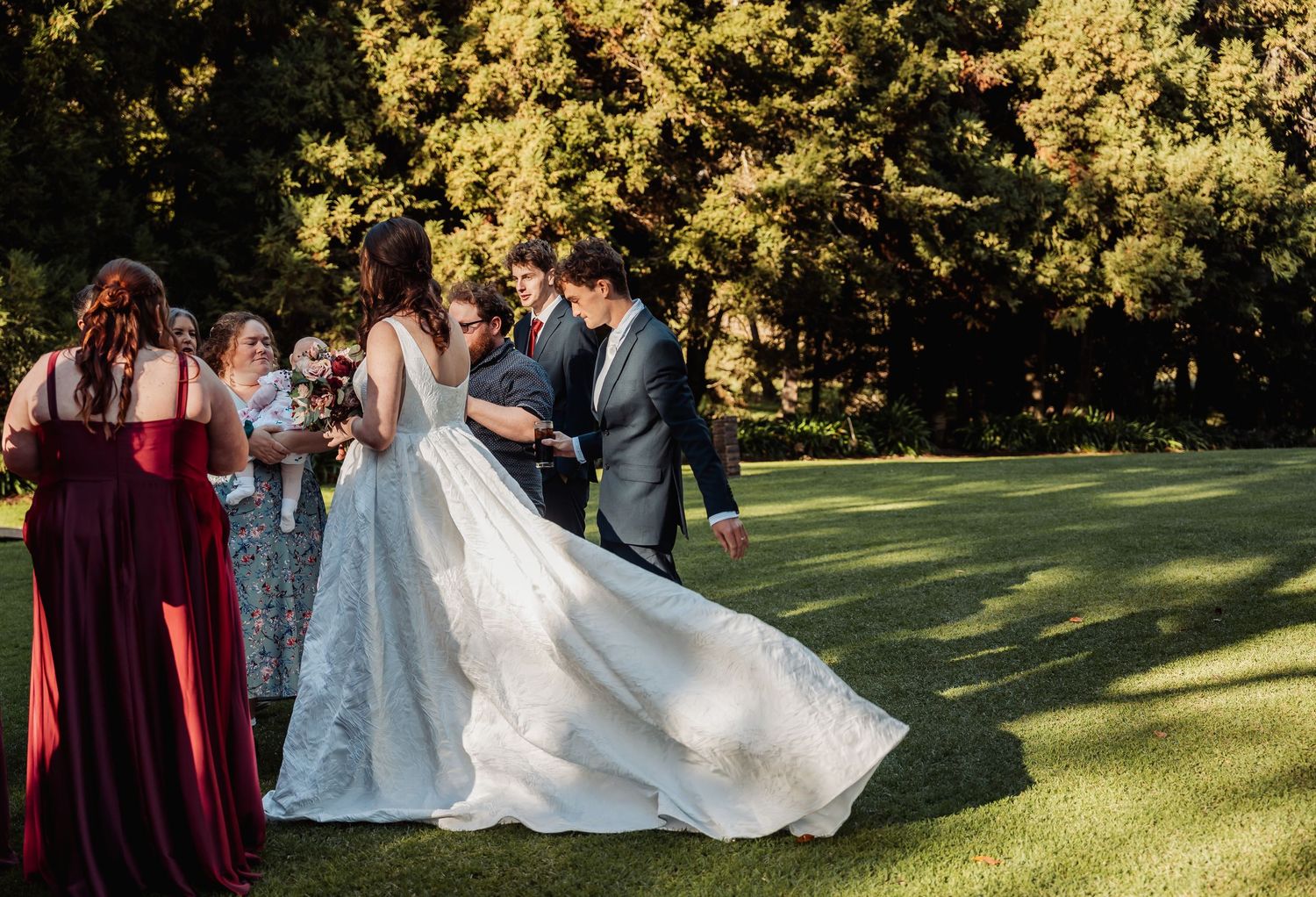 Wedding party helping bride with lace gown train on sunlit green lawn.