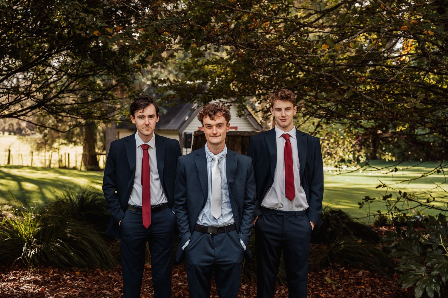 Three groomsmen in navy suits with red ties posing under tree canopy at outdoor wedding venue.