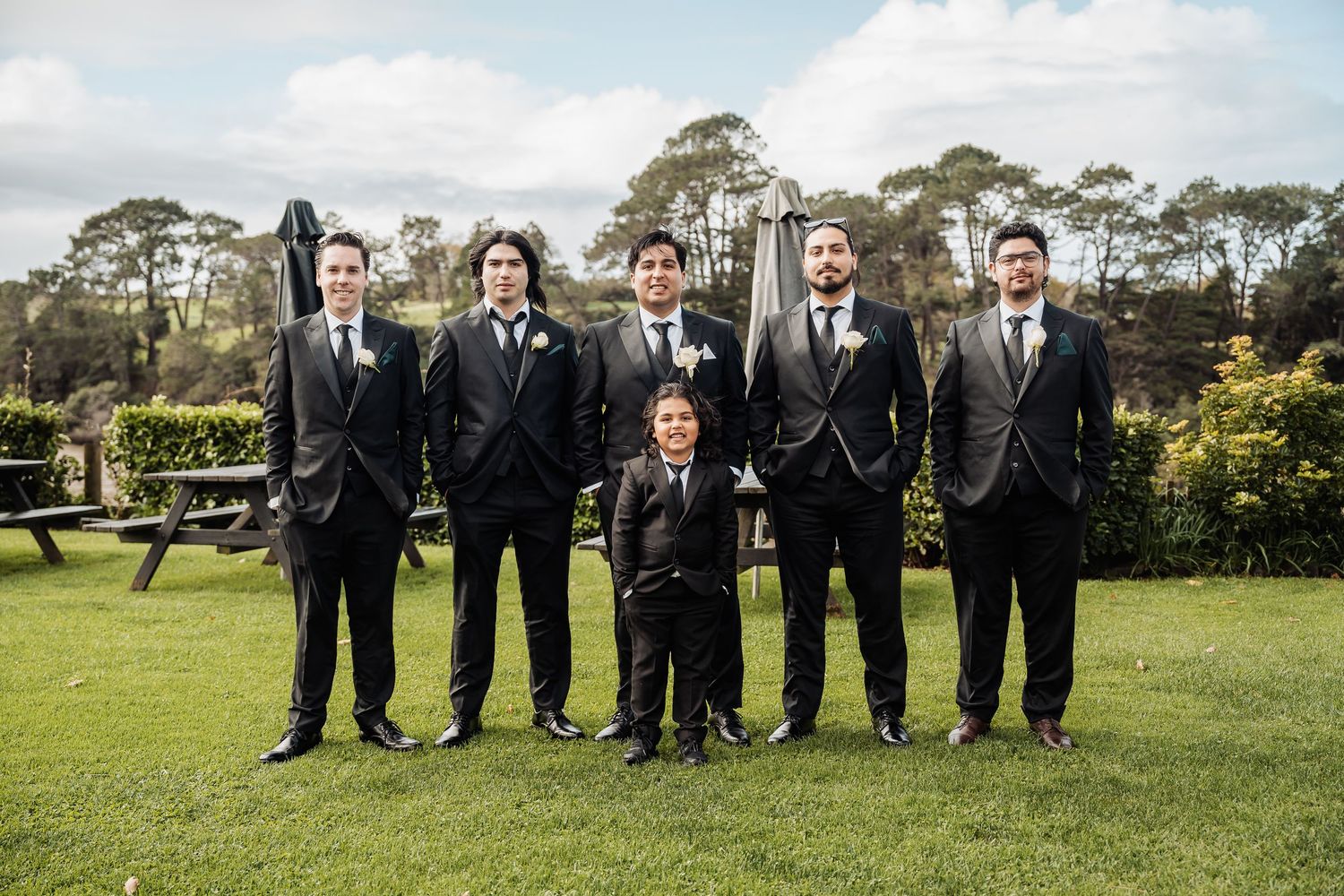A group stands together on a green lawn wearing matching black suits and ties with trees in the background.
