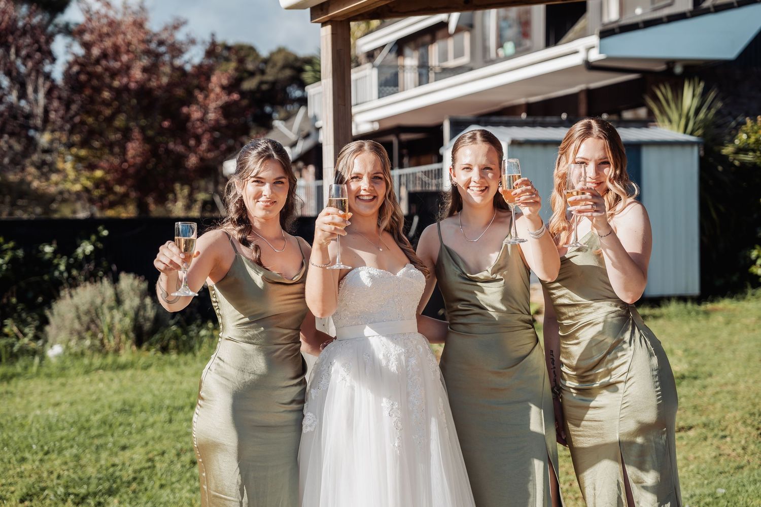 Group of people in sage green bridesmaid dresses pose with bride in white gown outdoors at a sunny wedding celebration.