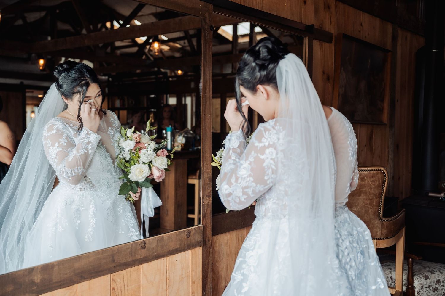 A bride in an elegant lace wedding gown and veil looks at her reflection in a rustic wooden mirror.