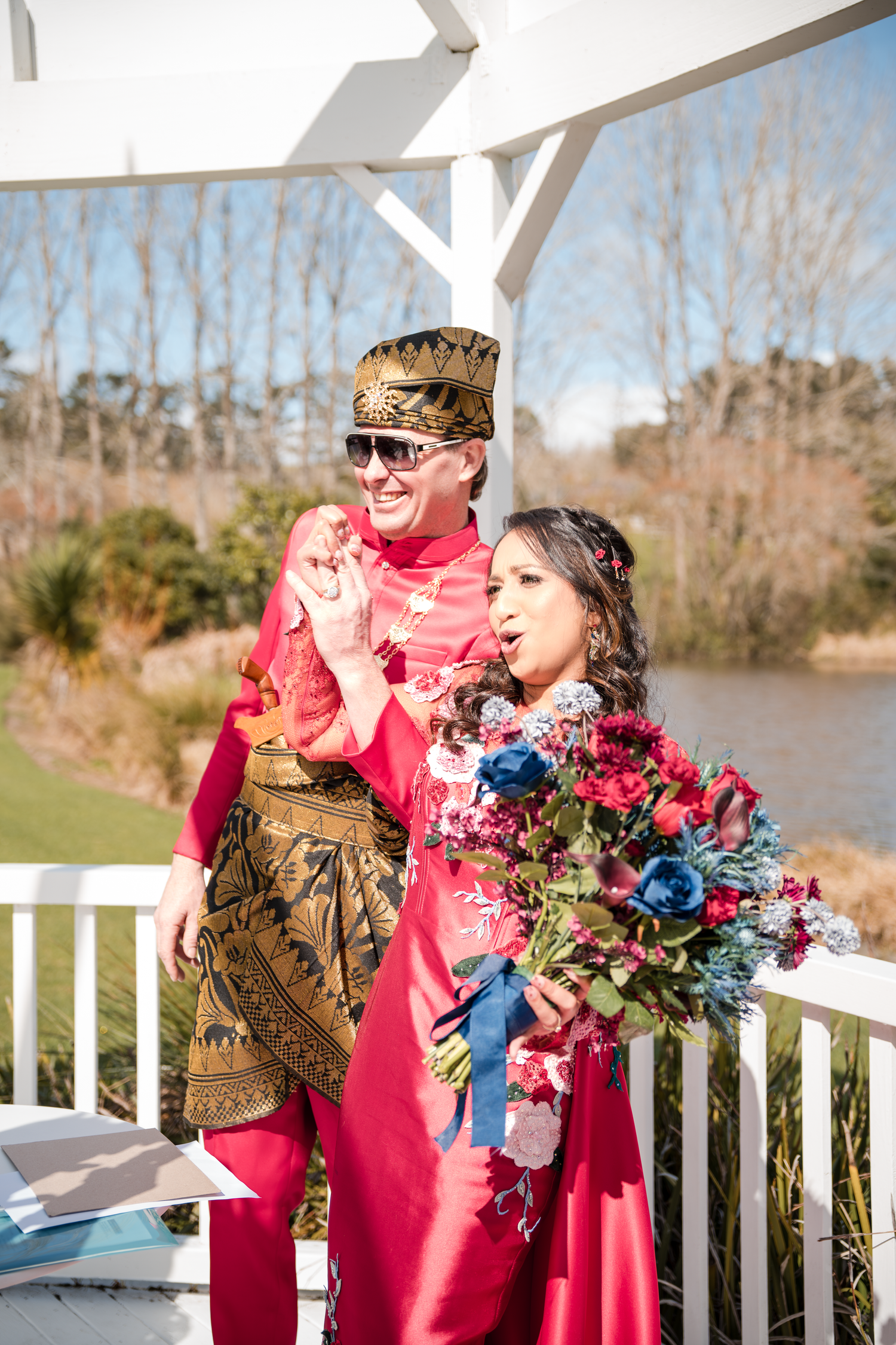 Two wedding party members in bright red attire pose on a white deck with a vibrant floral bouquet.