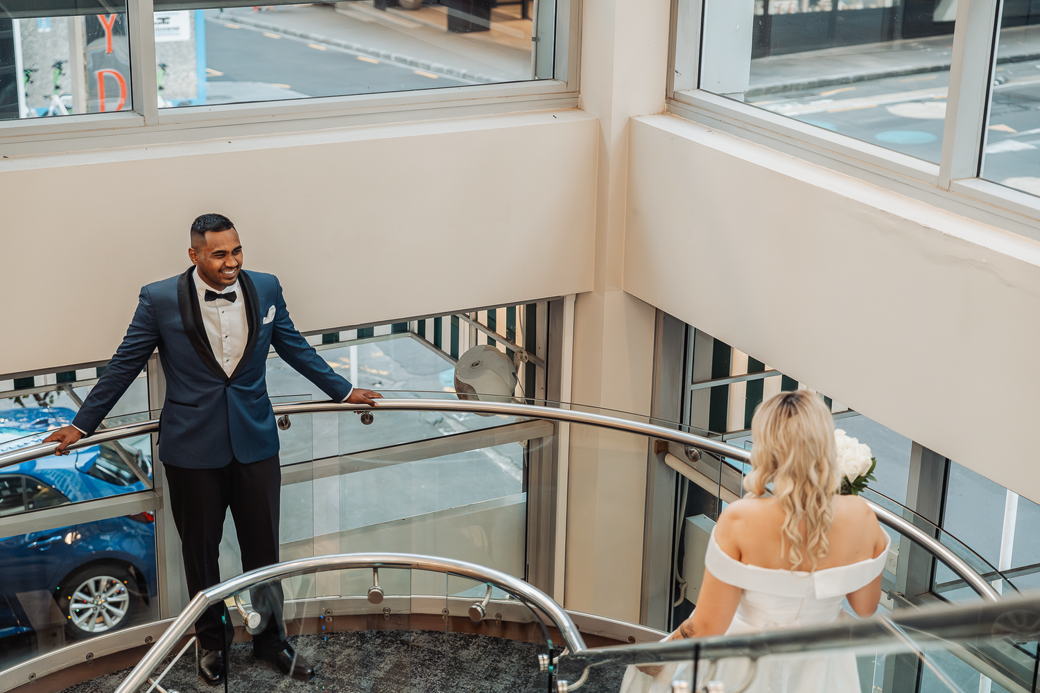 Wedding couple interacts playfully on a modern staircase with glass railings and natural lighting.