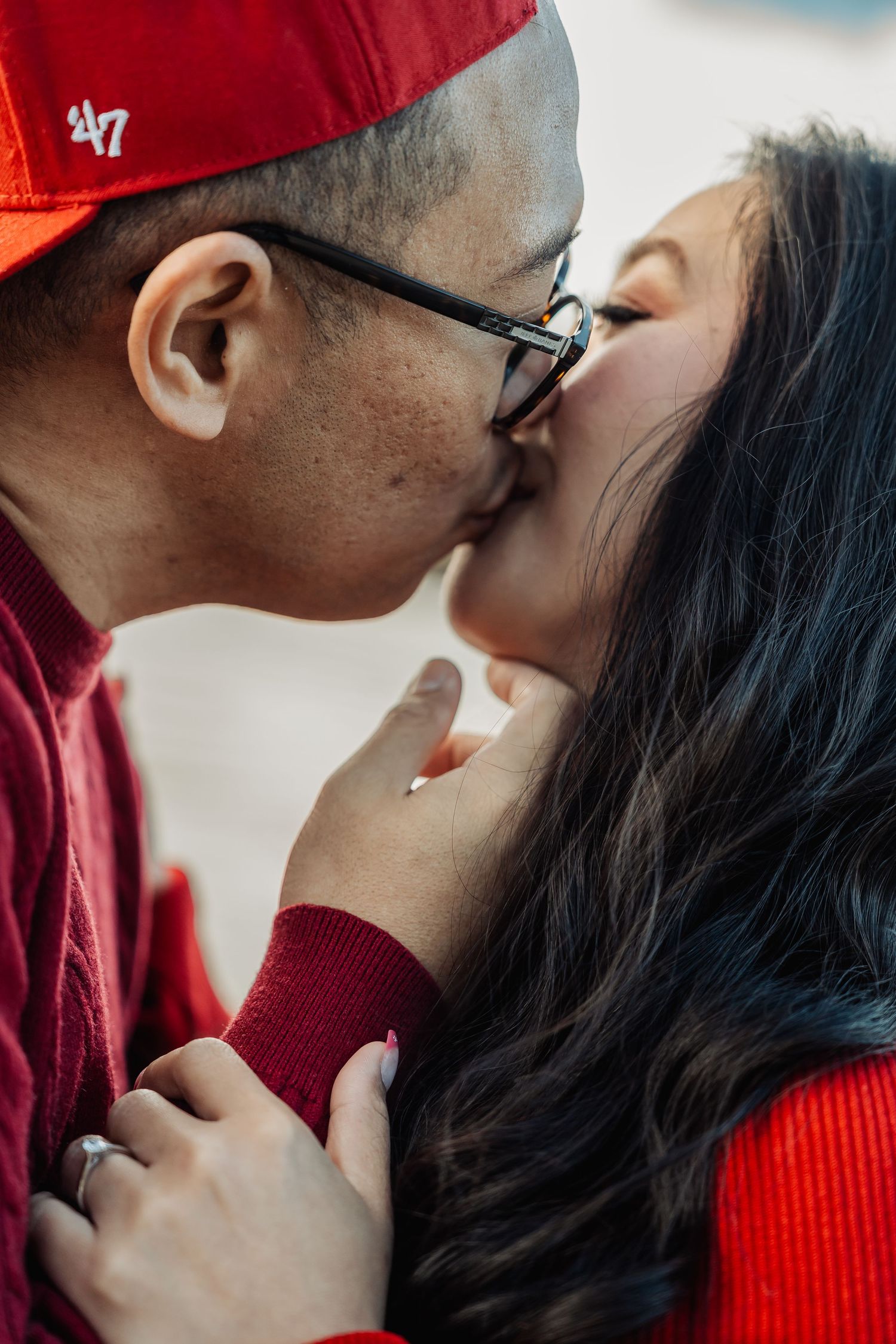 Close-up of a couple sharing a romantic kiss, man wearing red cap and glasses, woman in red dress.