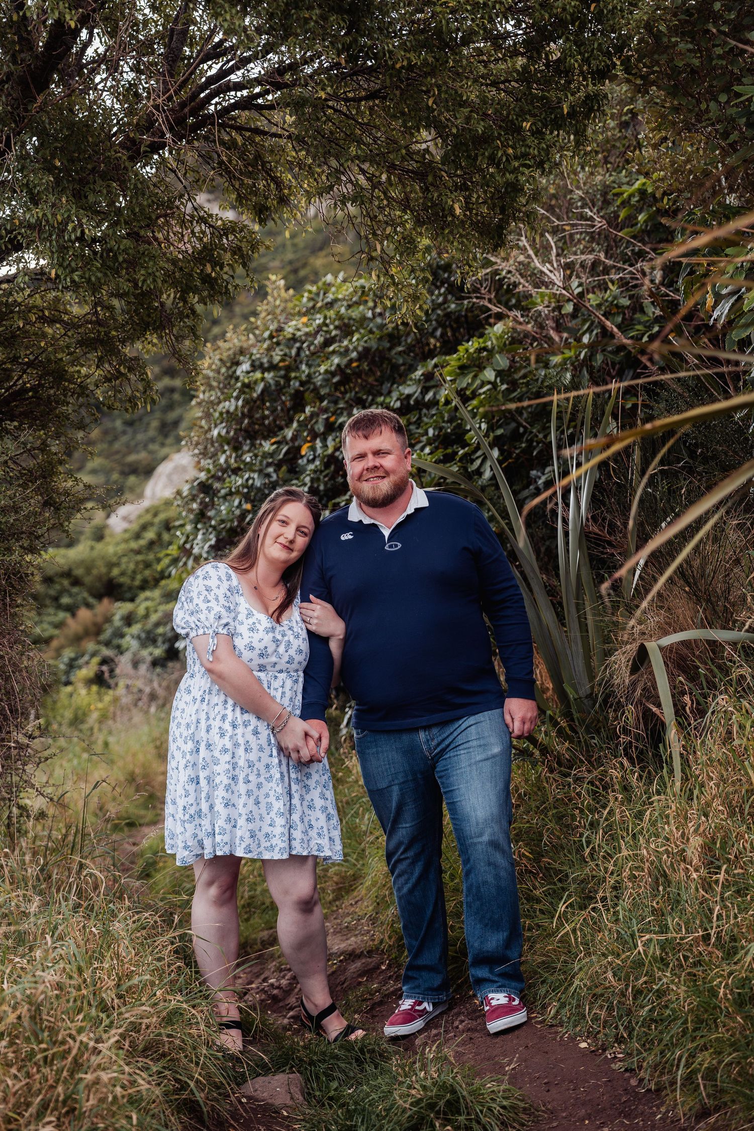 Couple holding hands on a lush forest trail, woman in floral dress and man in navy polo smiling at camera.
