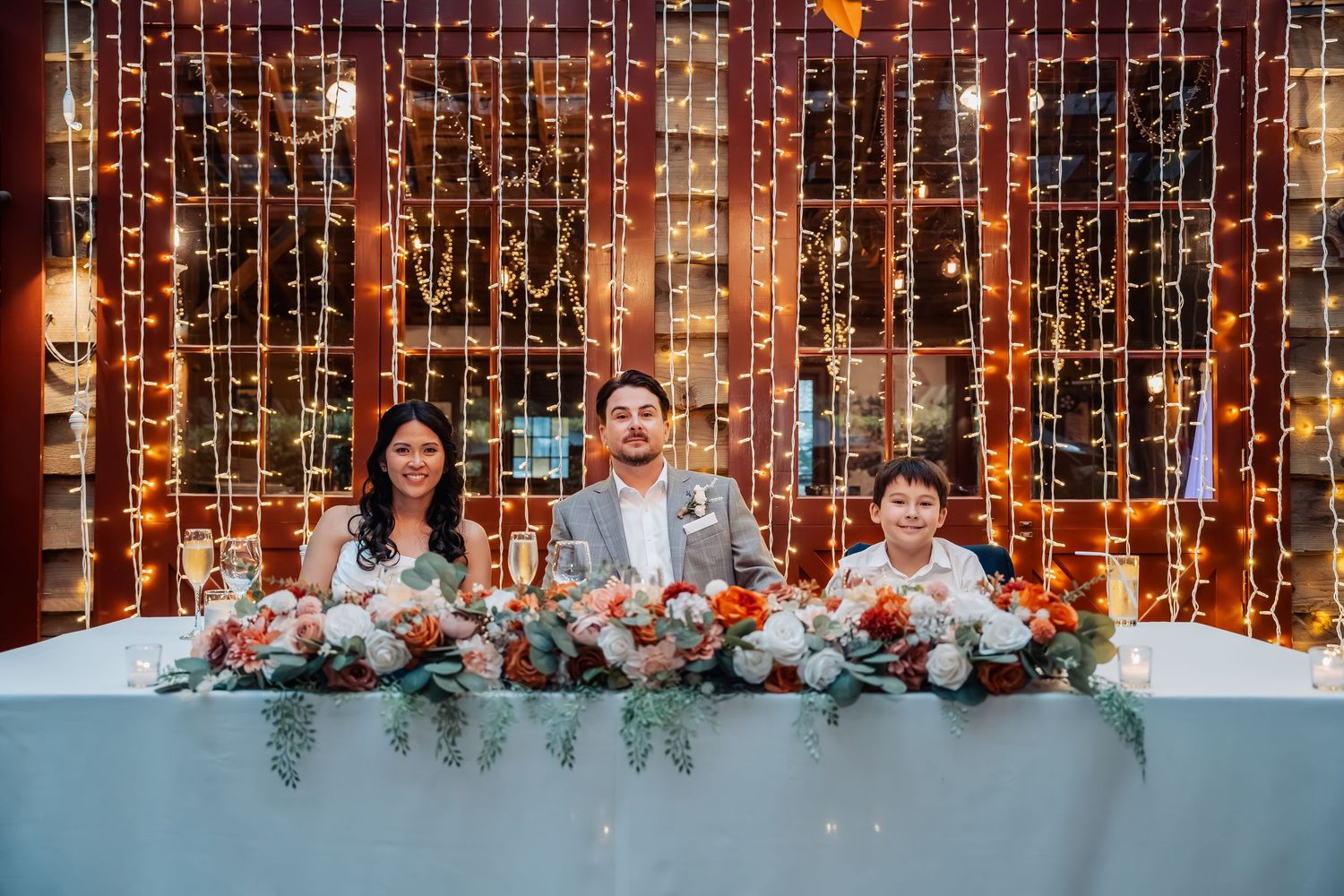Bride, groom, and child smile at sweetheart table adorned with floral arrangements and warm fairy light backdrop.