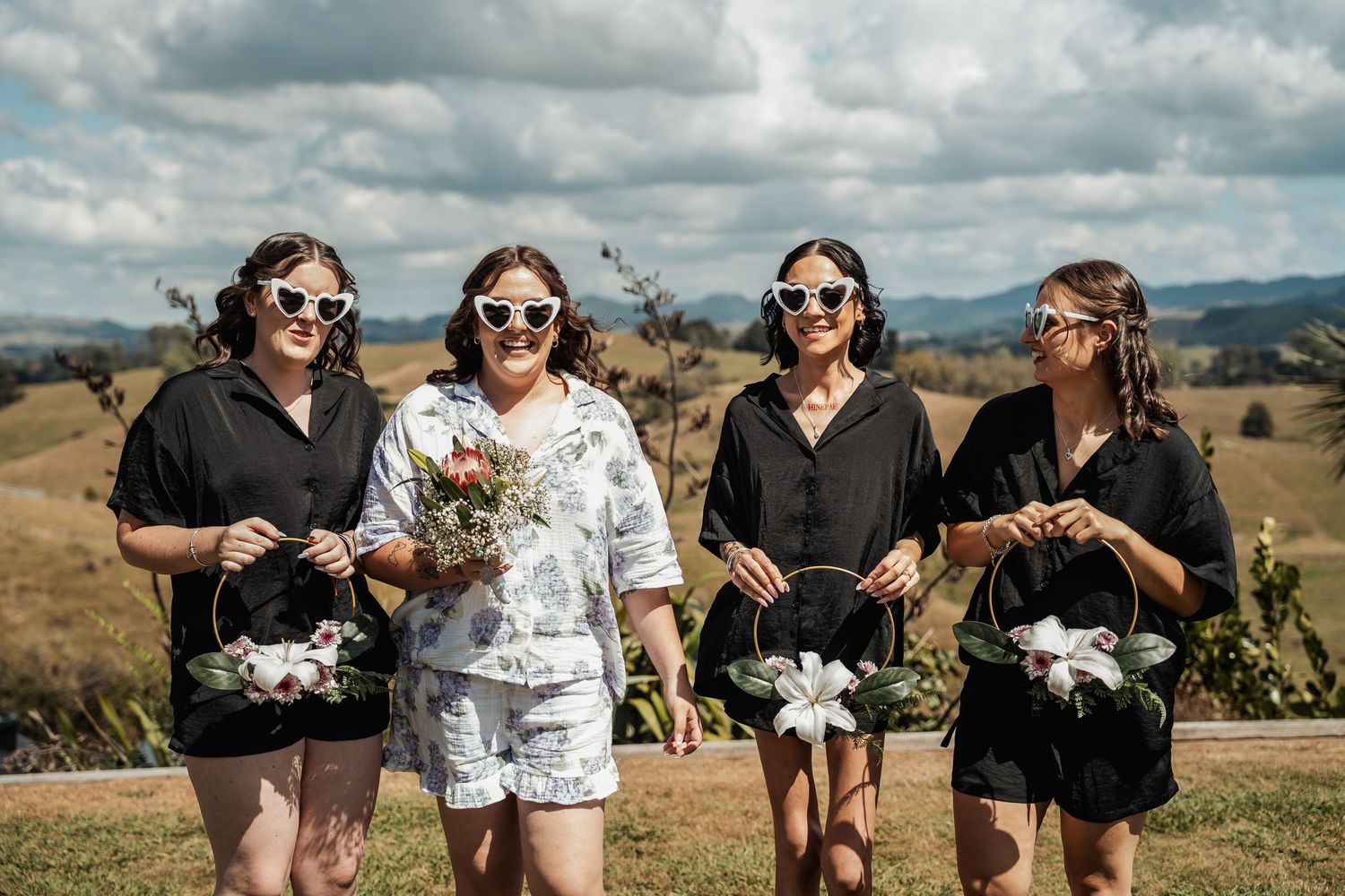 Four bridesmaids in black outfits wearing heart sunglasses holding bouquets in a scenic outdoor setting.