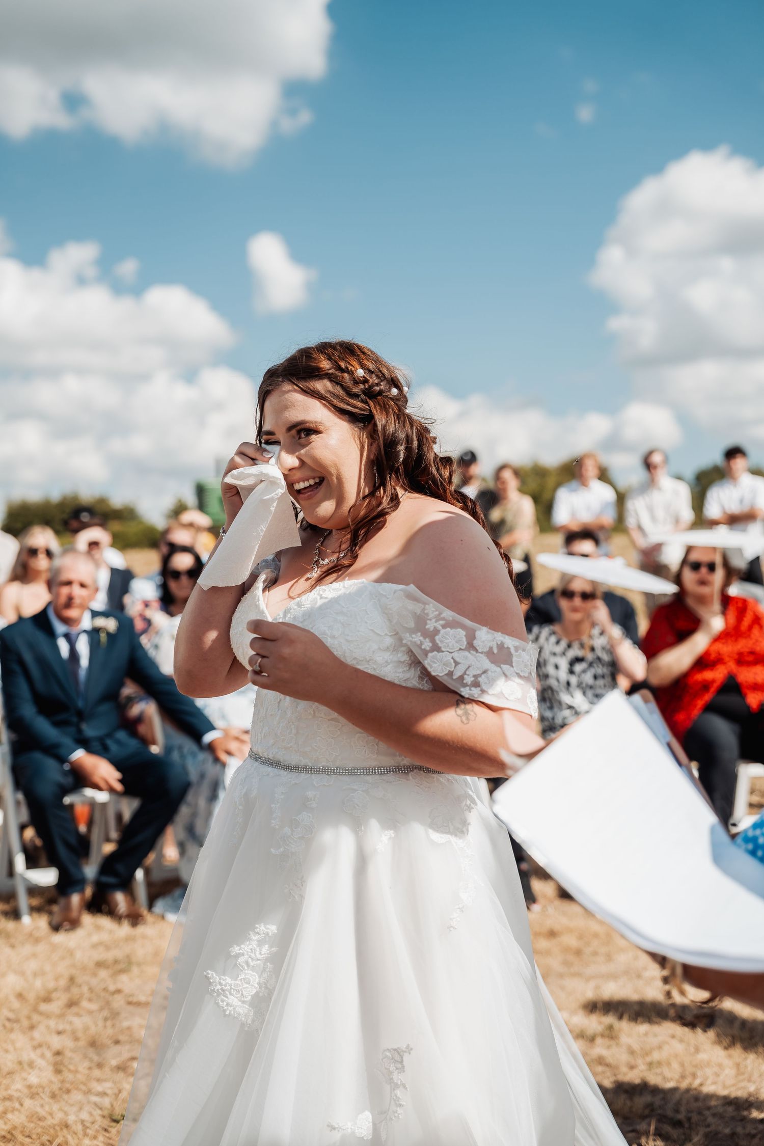 Emotional bride in off-shoulder lace gown wiping tears during outdoor wedding ceremony.