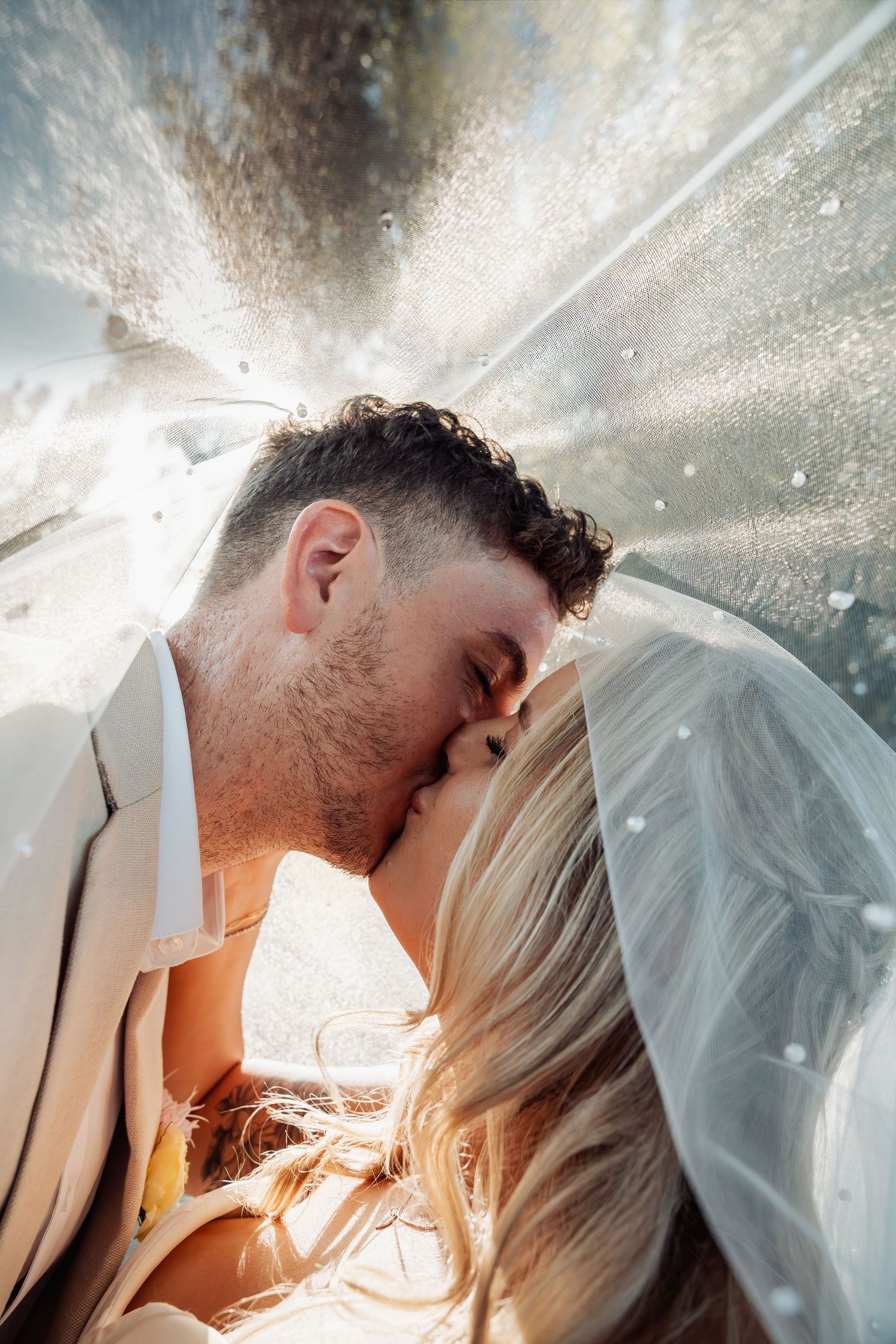 Bride and groom share a romantic kiss under a flowing wedding veil with sunlight streaming through.