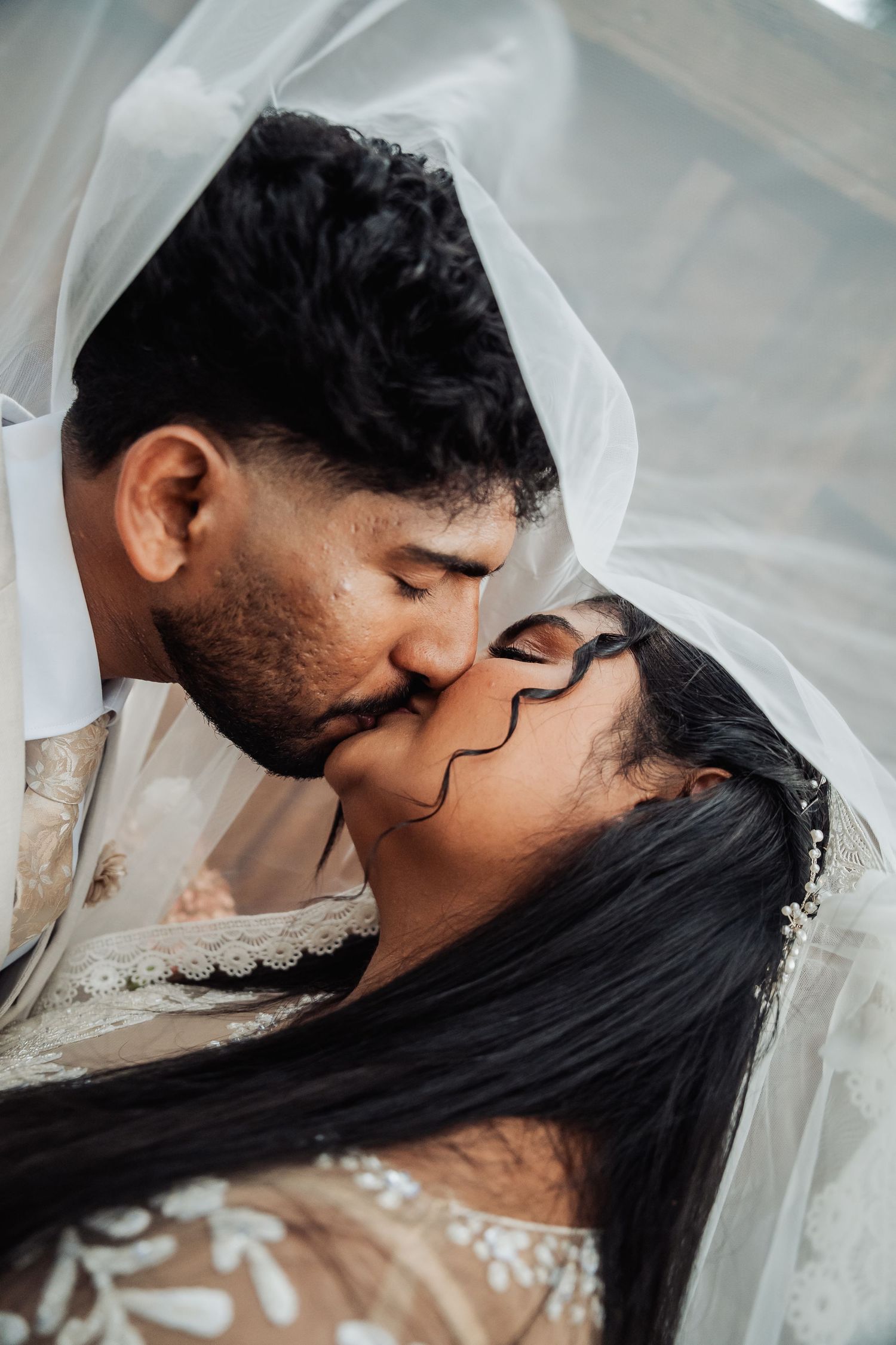 Bride and groom sharing a romantic kiss under a flowing white veil on their wedding day.