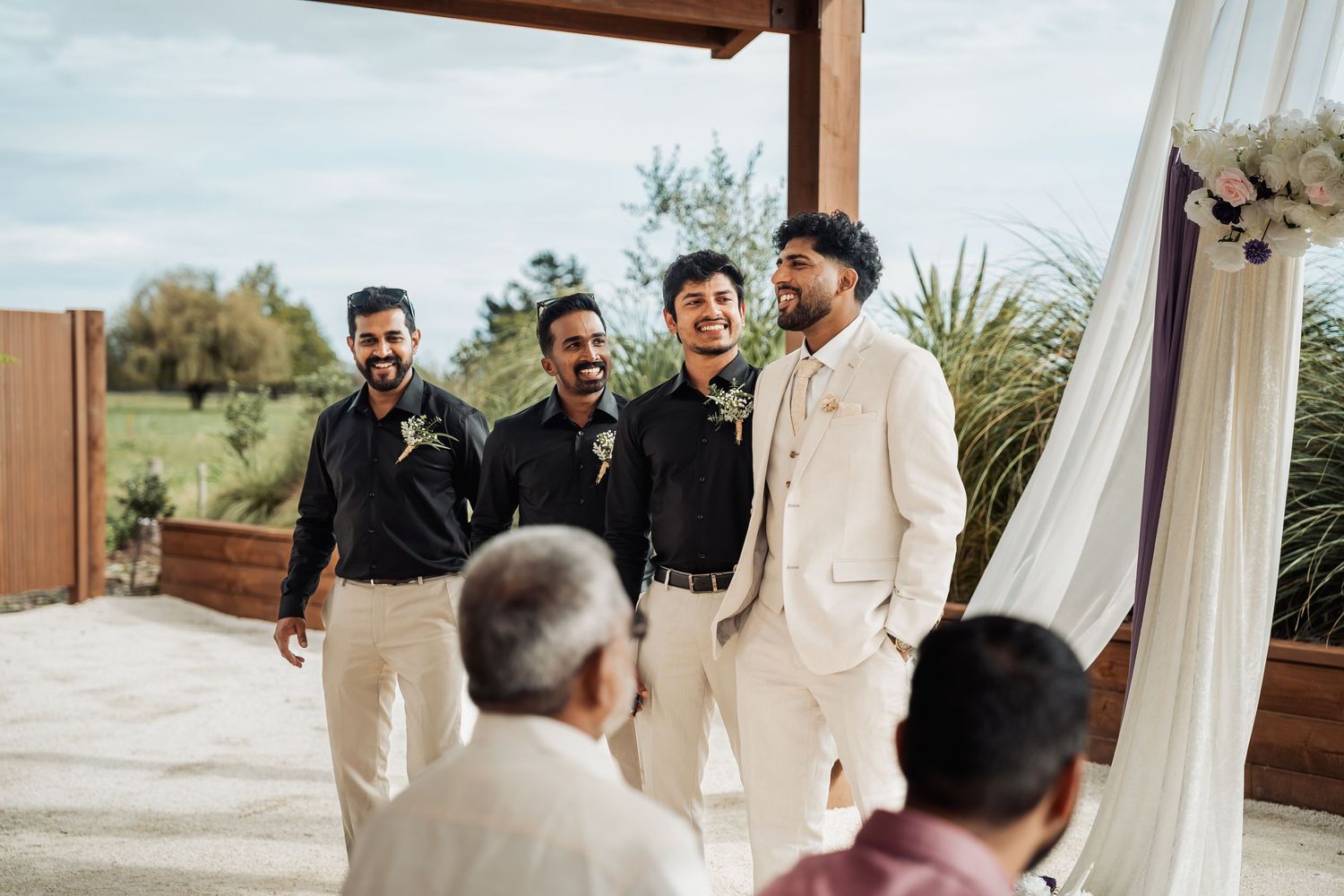 Groom in ivory suit laughing with groomsmen in black at outdoor wedding ceremony.