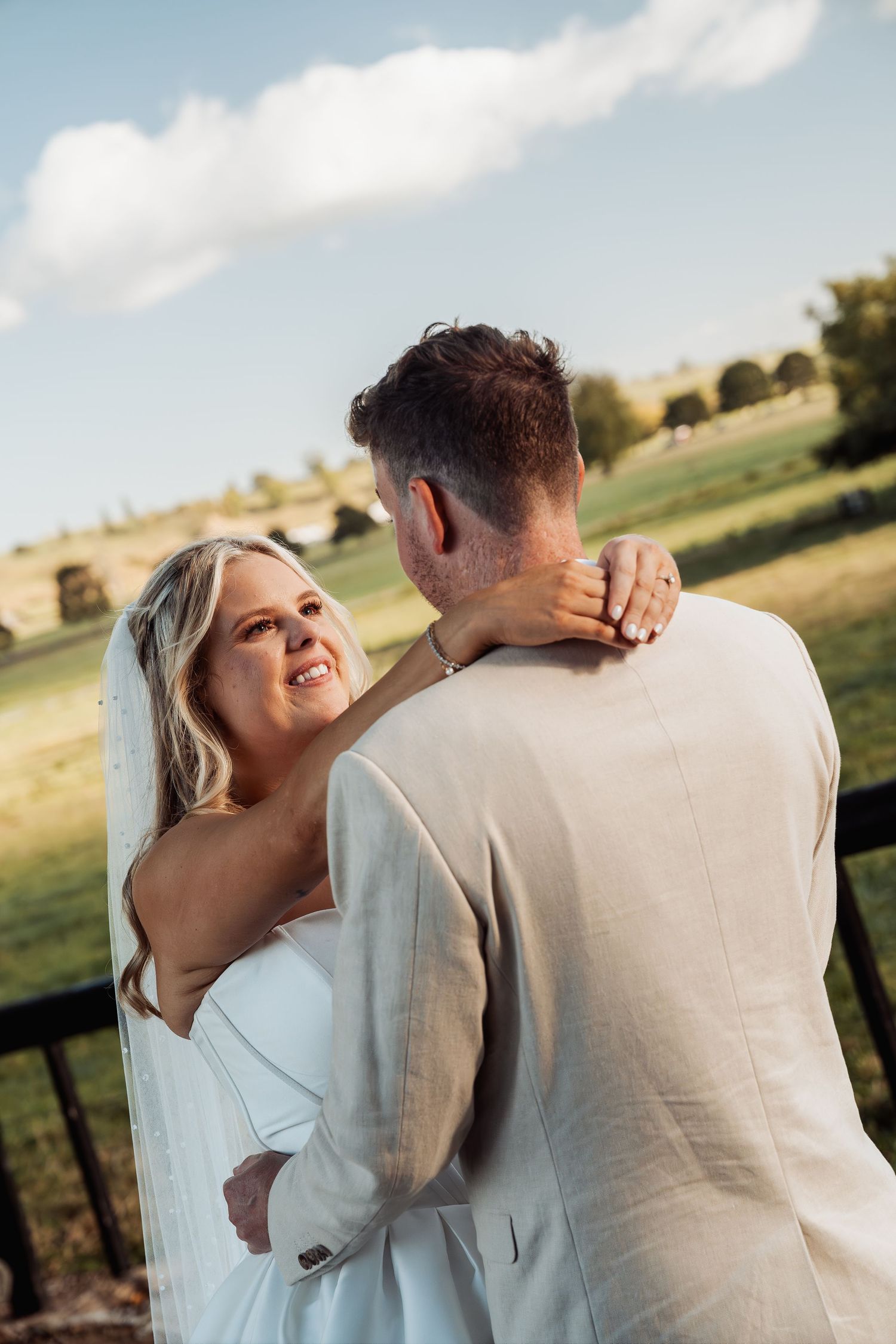 A glowing bride in a white gown and veil smiles at her groom against a scenic green countryside backdrop.