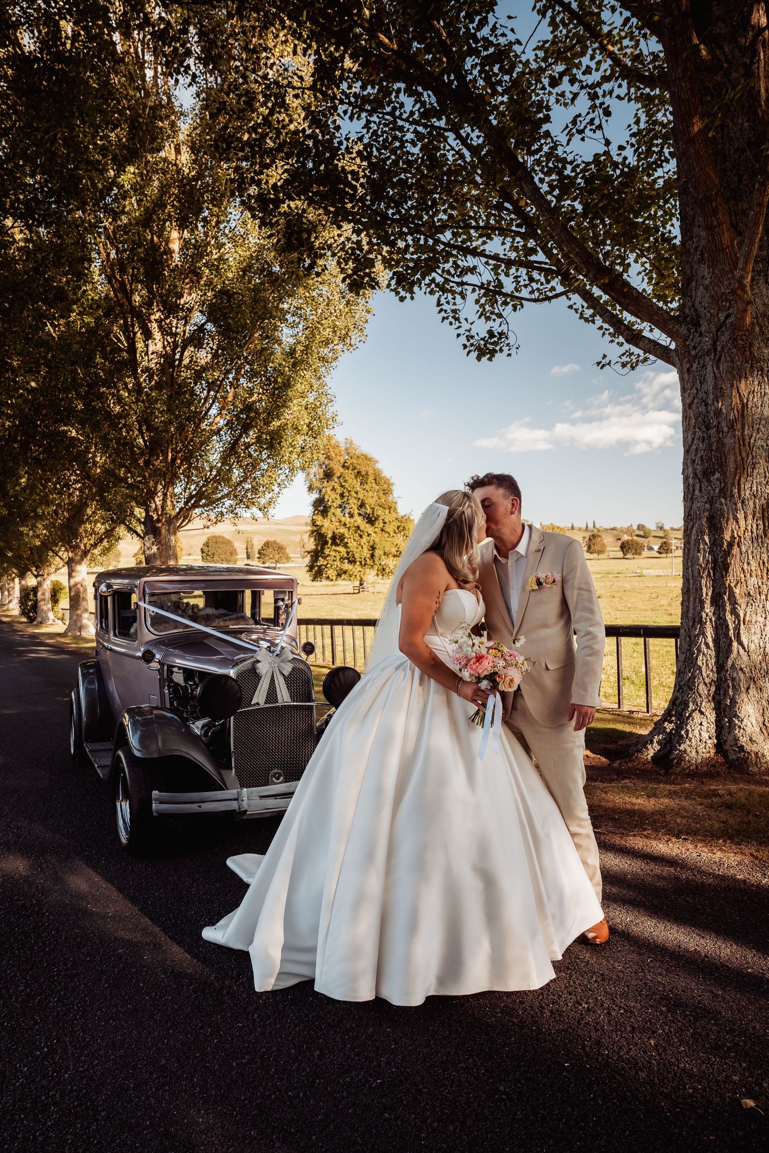 Bride and groom kissing beside a vintage classic car on a tree-lined driveway on their wedding day.