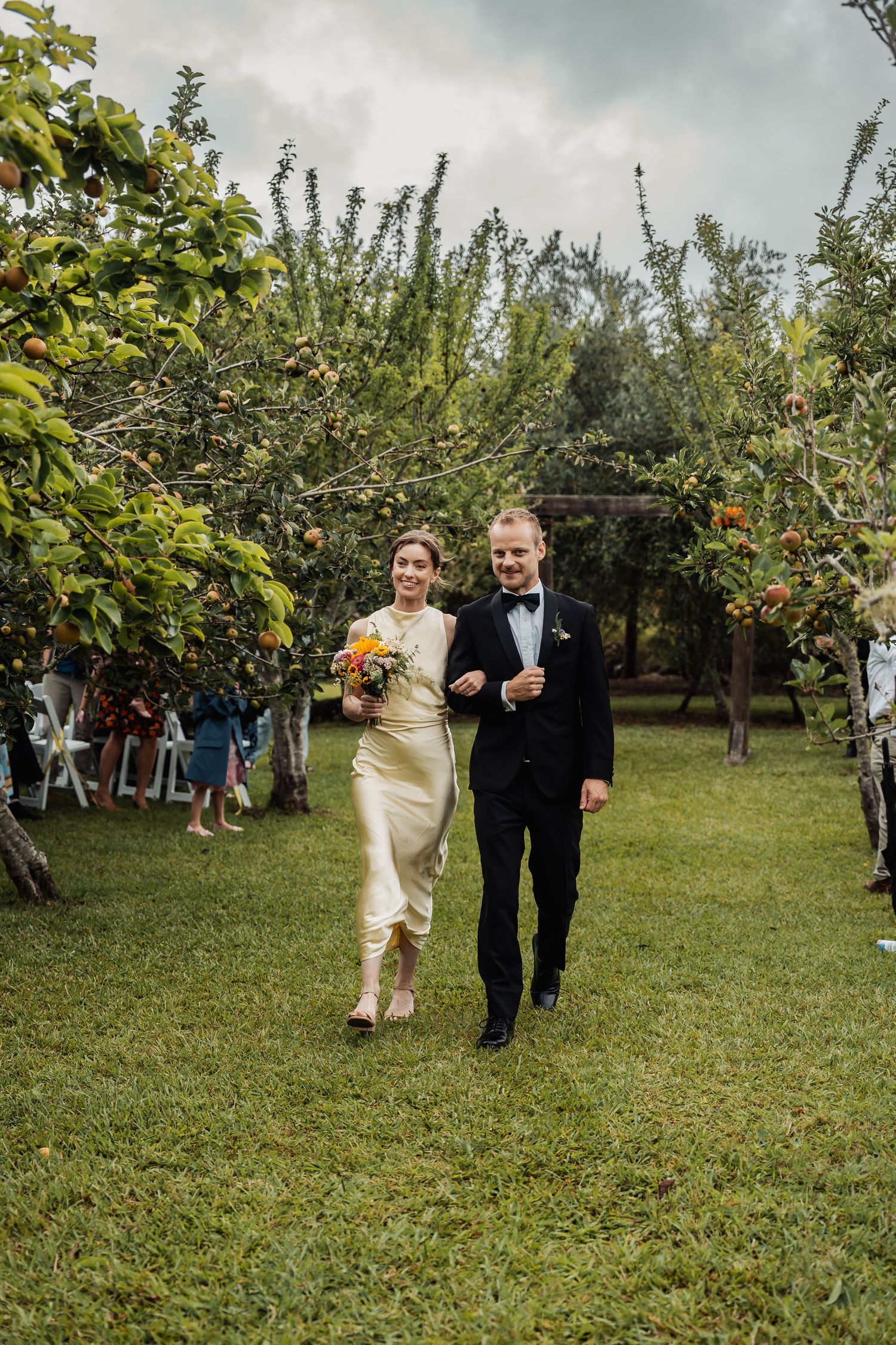 Smiling newlyweds walk arm in arm down the aisle through a lush orchard after their outdoor garden wedding ceremony.
