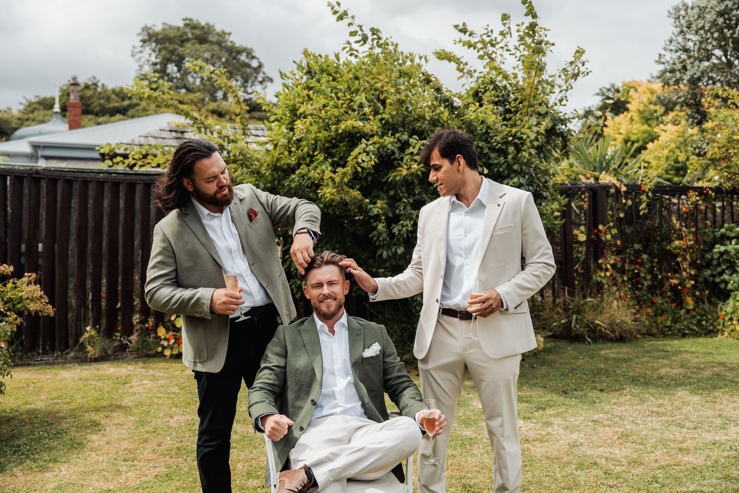 Three groomsmen laugh together in a garden, one seated in a chair, wearing suits for a wedding.