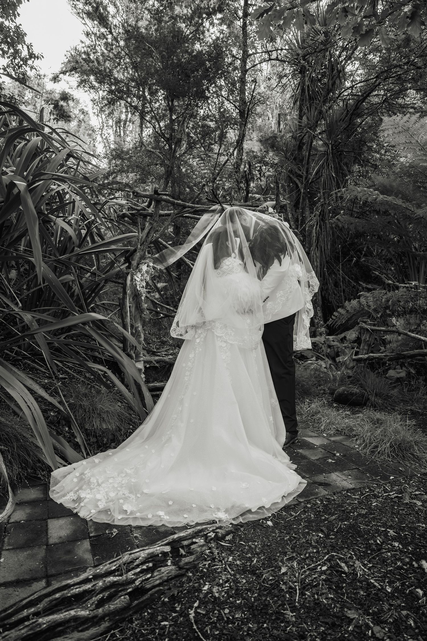 Black and white photo of bride and groom kissing under flowing veil in lush forest setting.