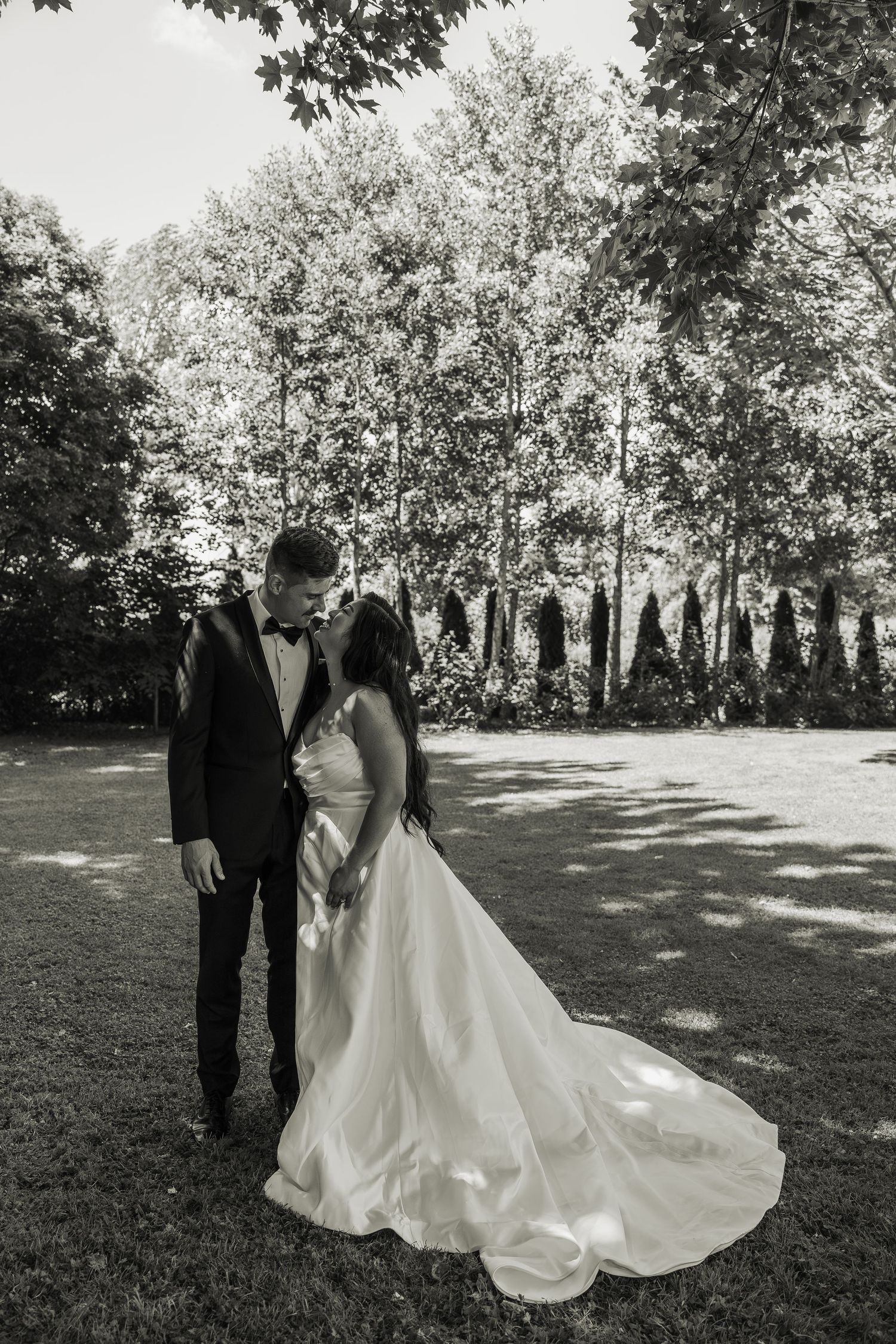 Black and white portrait of bride and groom standing in sunlit tree-lined garden.