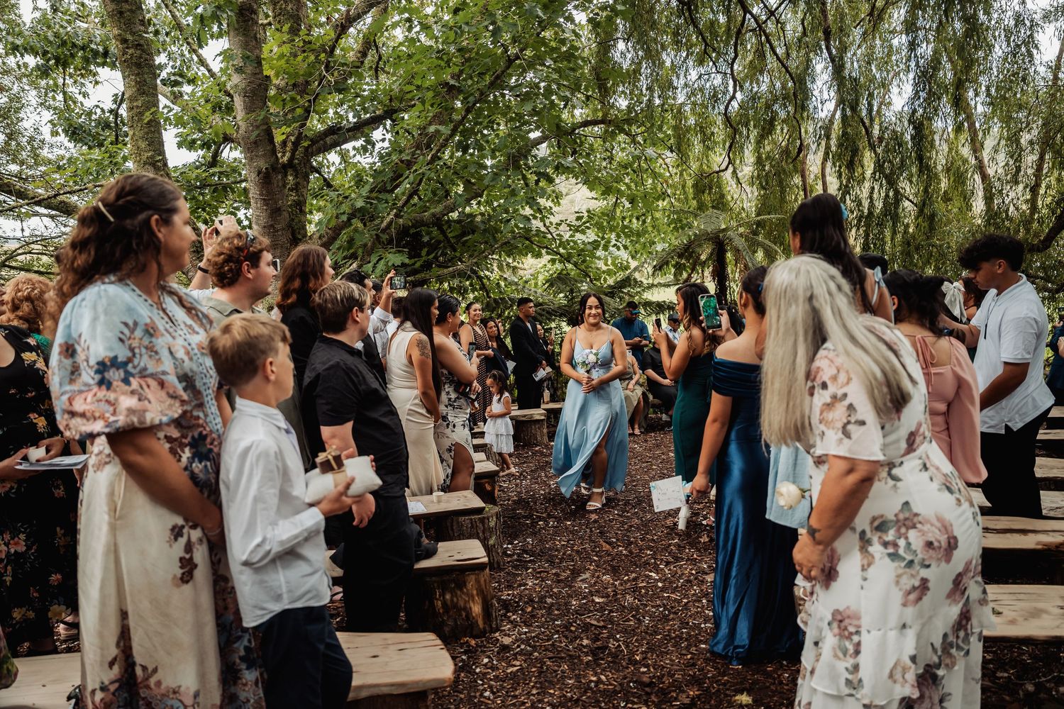 Outdoor woodland wedding ceremony with guests standing among rustic benches, surrounded by lush green trees.