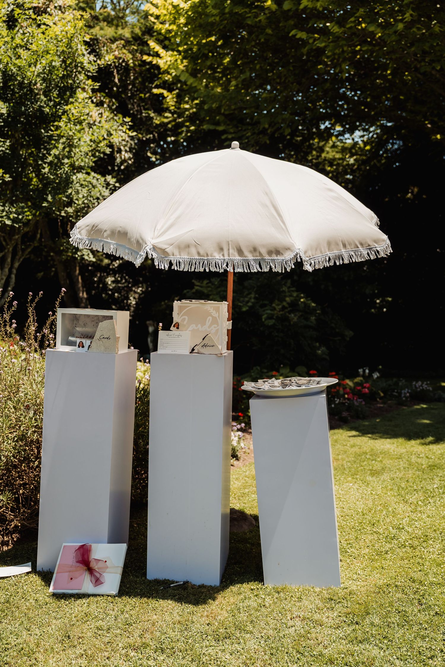 White fringed patio umbrella over elegant white pedestals with wedding gifts on green lawn.