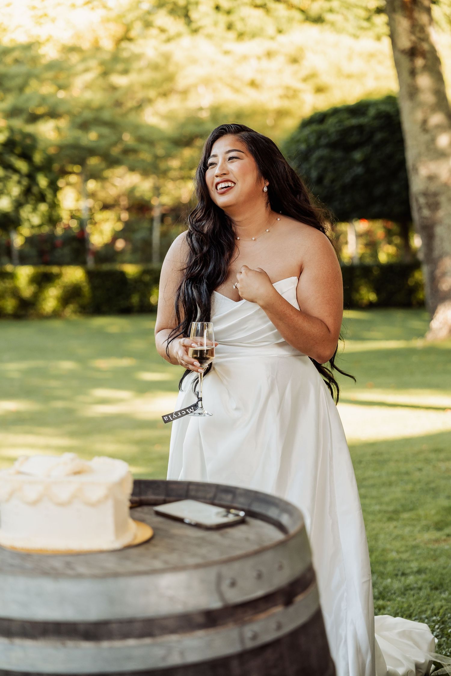 Bride in white strapless gown laughing outdoors holding wine glass near wedding cake on rustic barrel.