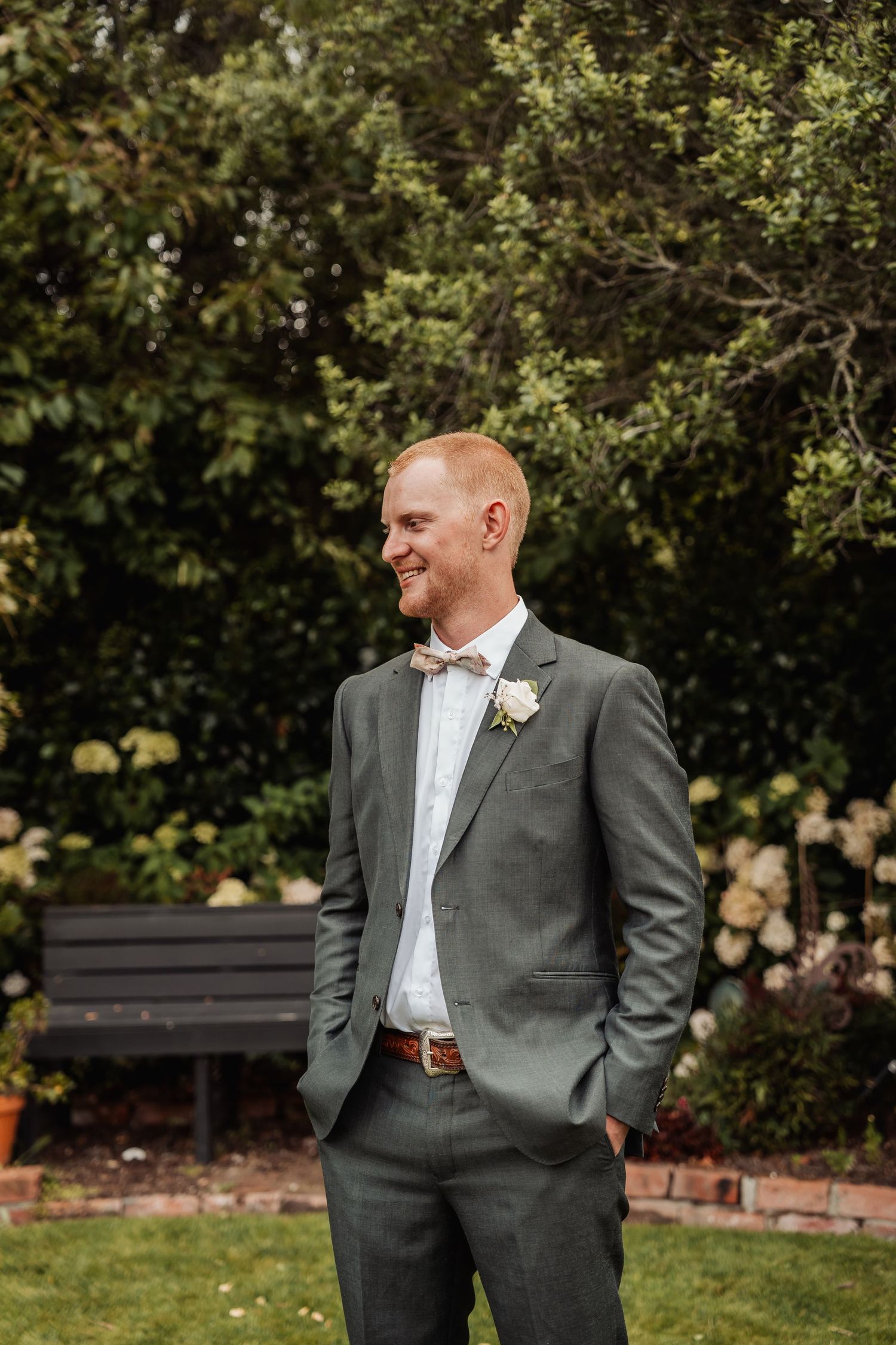 Smiling groom in grey suit with white boutonniere stands in a lush garden setting.